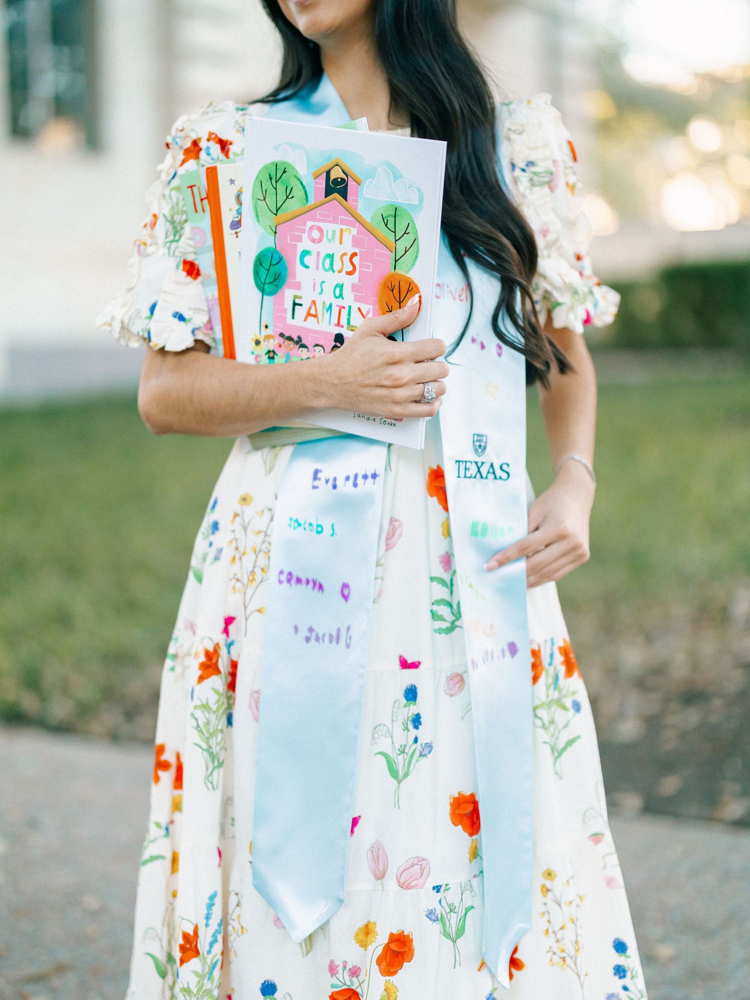 Woman in floral dress holding a Texas graduation sash and colorful class book at an outdoor ceremony.