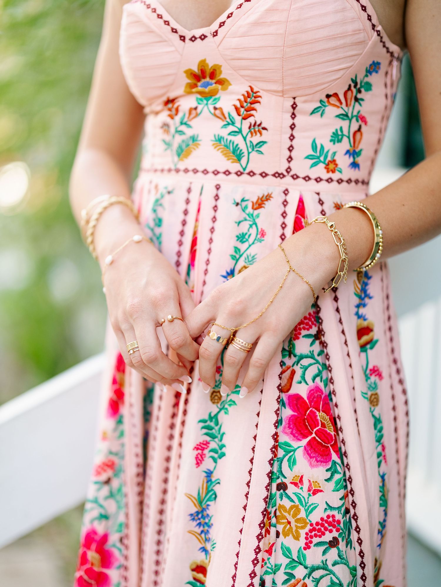 Woman wearing a pink floral embroidered maxi dress with gold rings and bracelets, showcasing intricate colorful needlework.