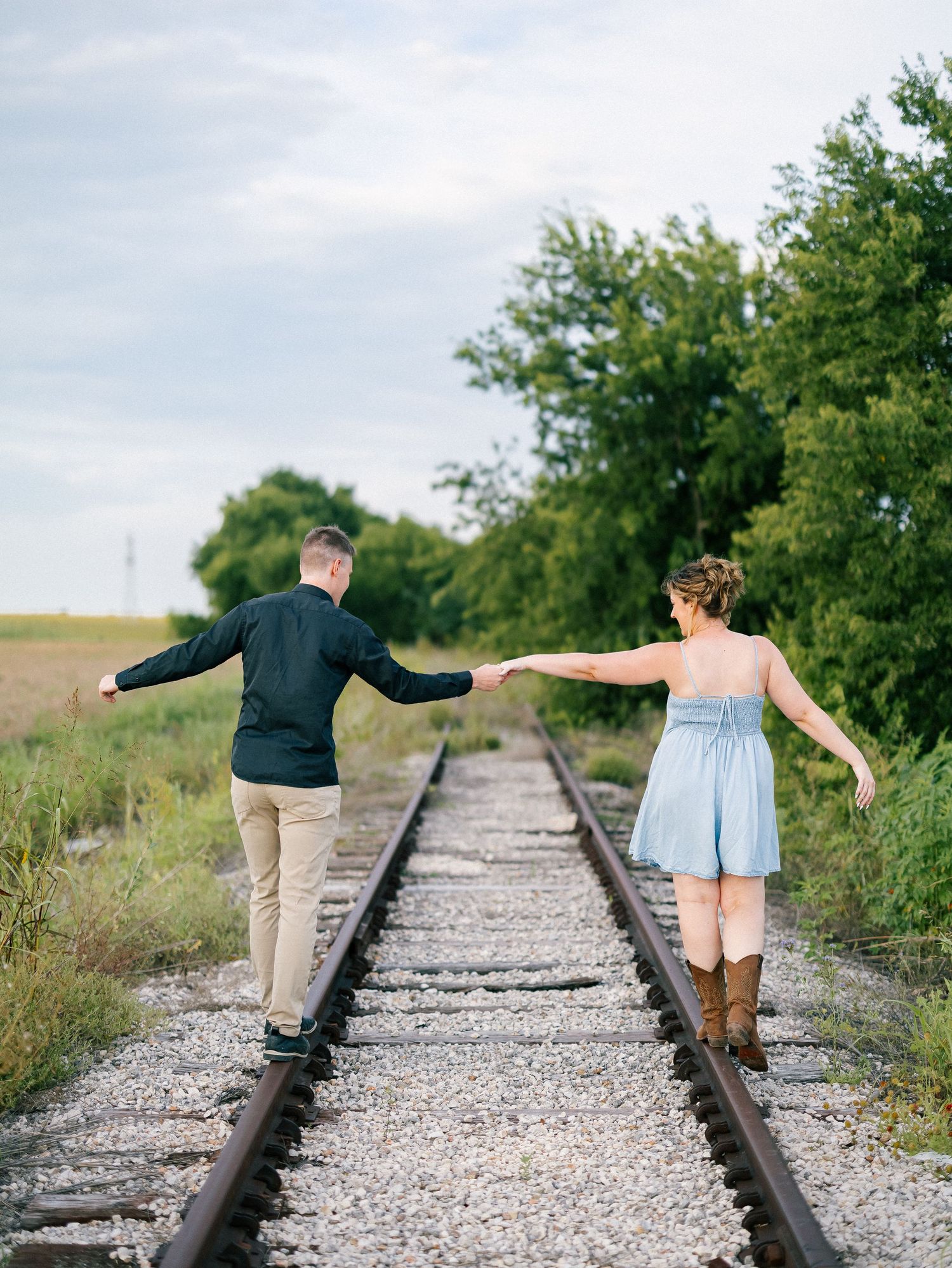 Couple holding hands while walking along railroad tracks through a scenic rural landscape.