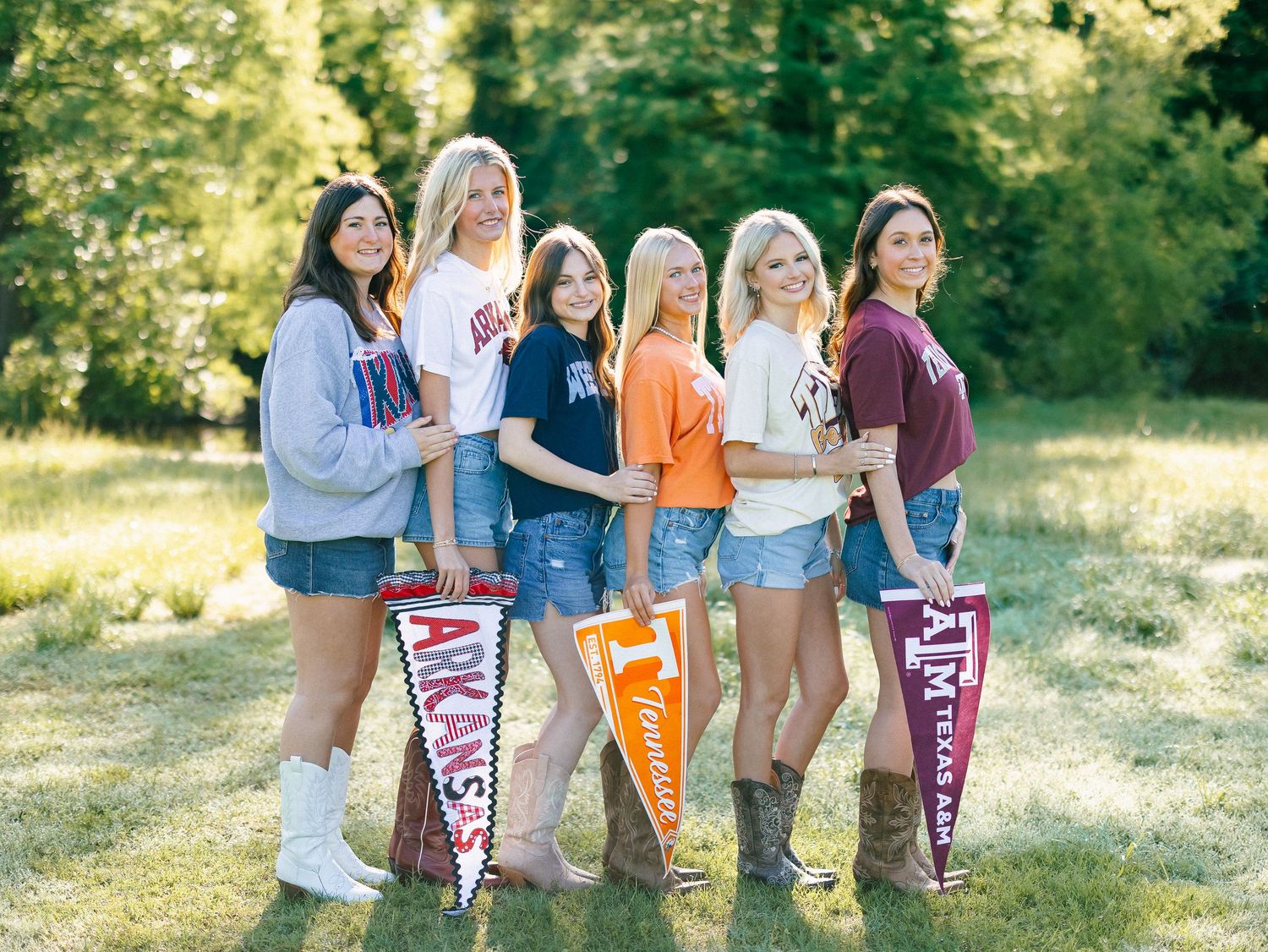 Six young women pose outdoors holding college pennants including Arkansas, Tennessee, and Texas A&M flags.