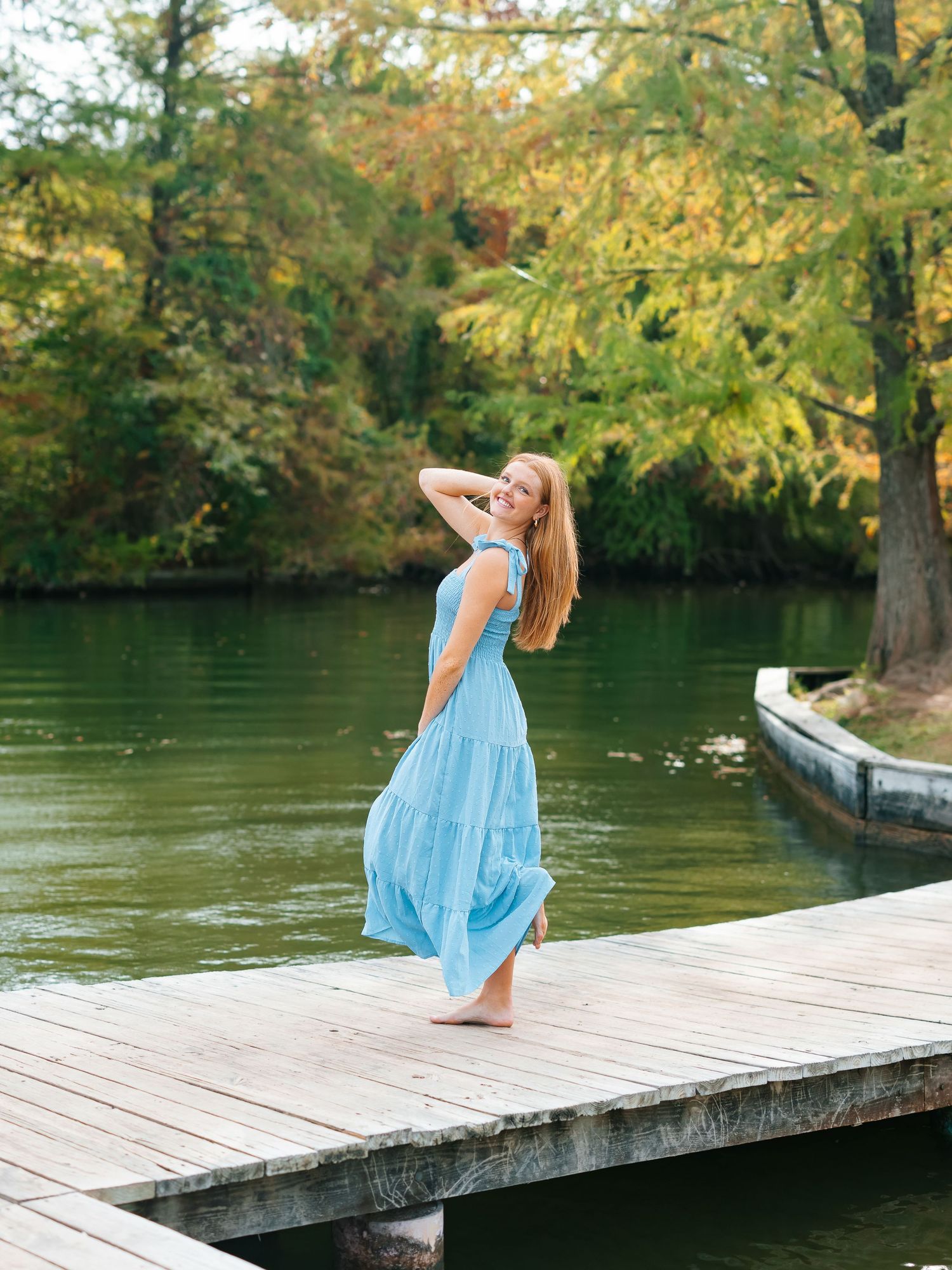 Young woman in a blue maxi dress posing on a wooden dock by an autumn lake.