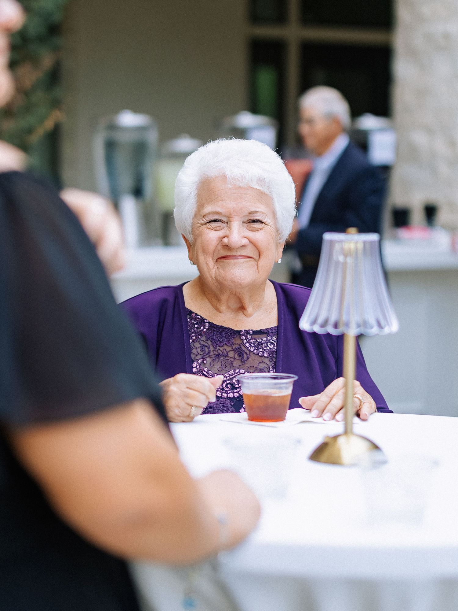Elderly woman in purple top smiling at outdoor event, holding a drink at a white table with a small lamp.