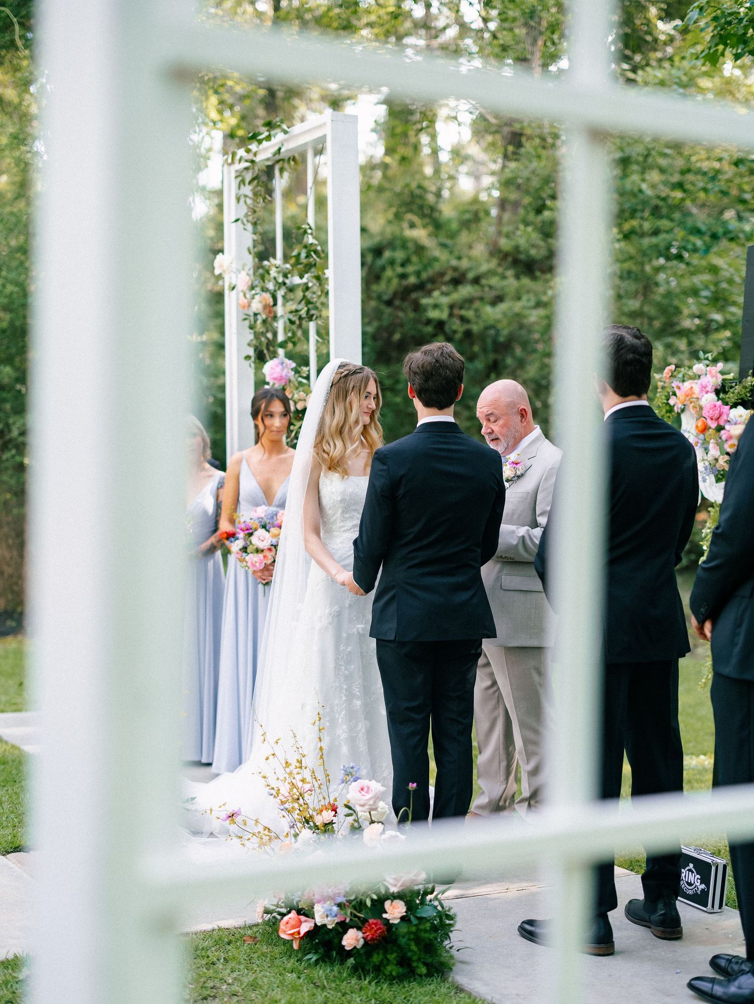 Outdoor wedding ceremony with bride in white lace gown, bridesmaids in blue, beneath a floral arch surrounded by lush greenery.