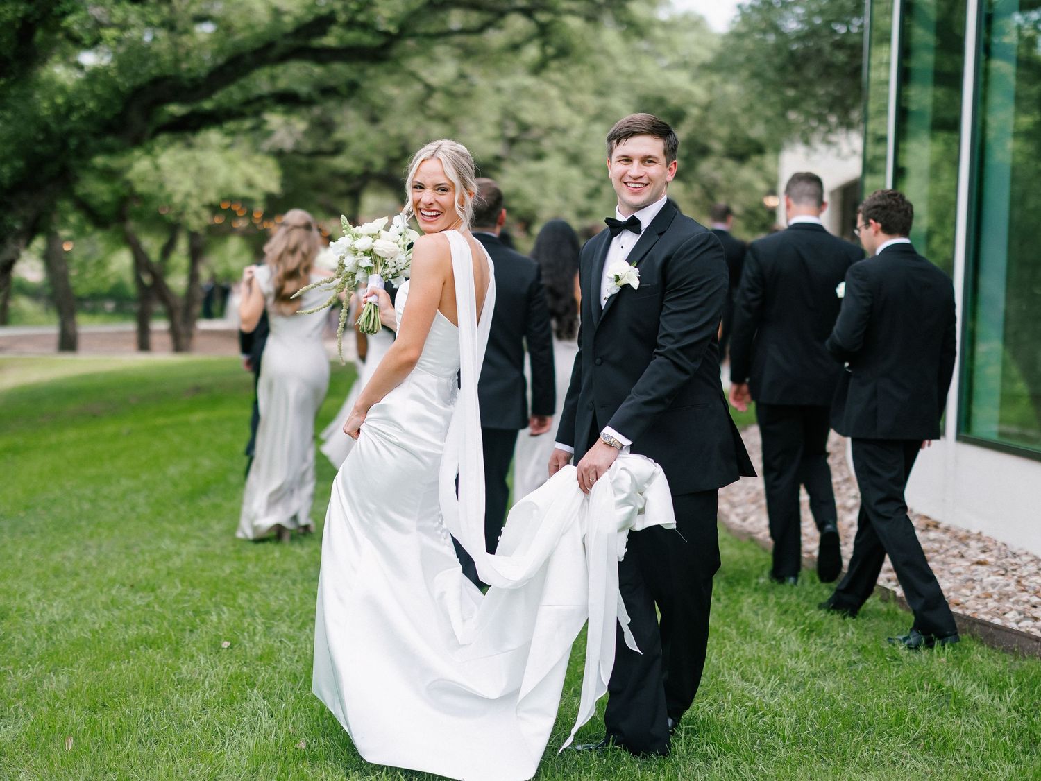 Bride and groom walk hand-in-hand on green lawn, wedding party behind them, bride in white gown holding bouquet.