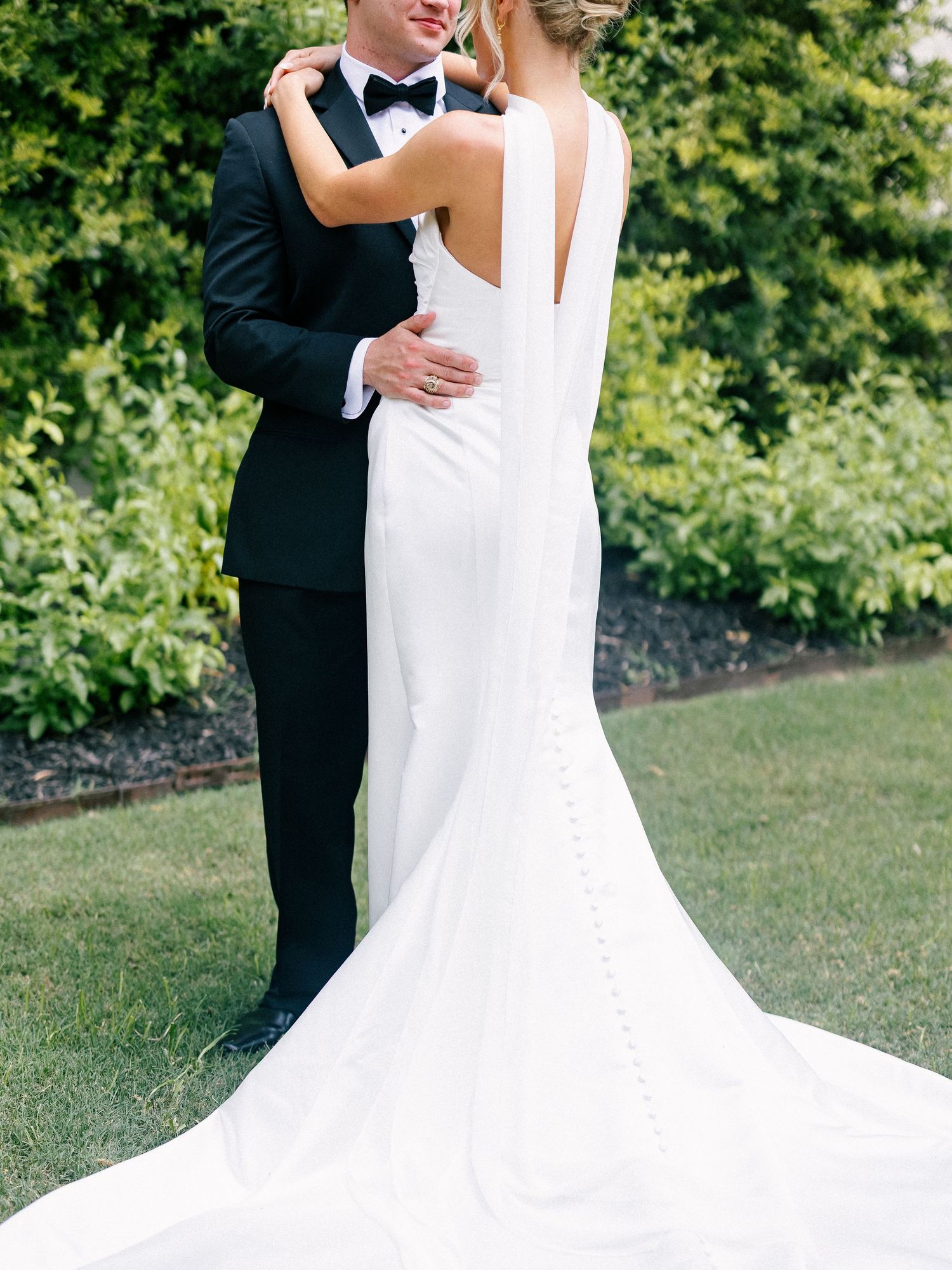 Bride in elegant white backless wedding gown with train embraces groom in black tuxedo outdoors.