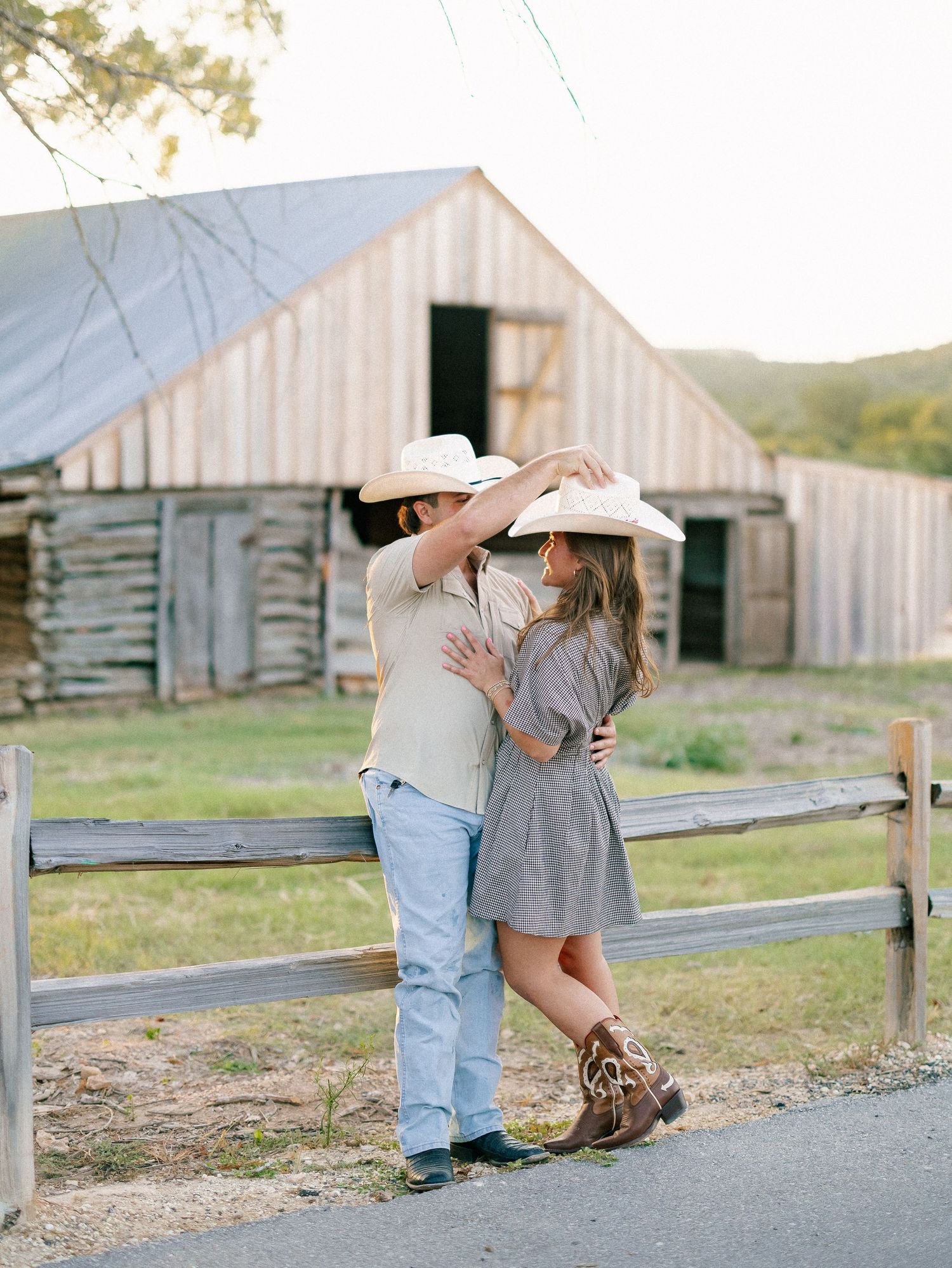 Couple in cowboy hats sharing a romantic moment by a rustic fence with a weathered barn backdrop.