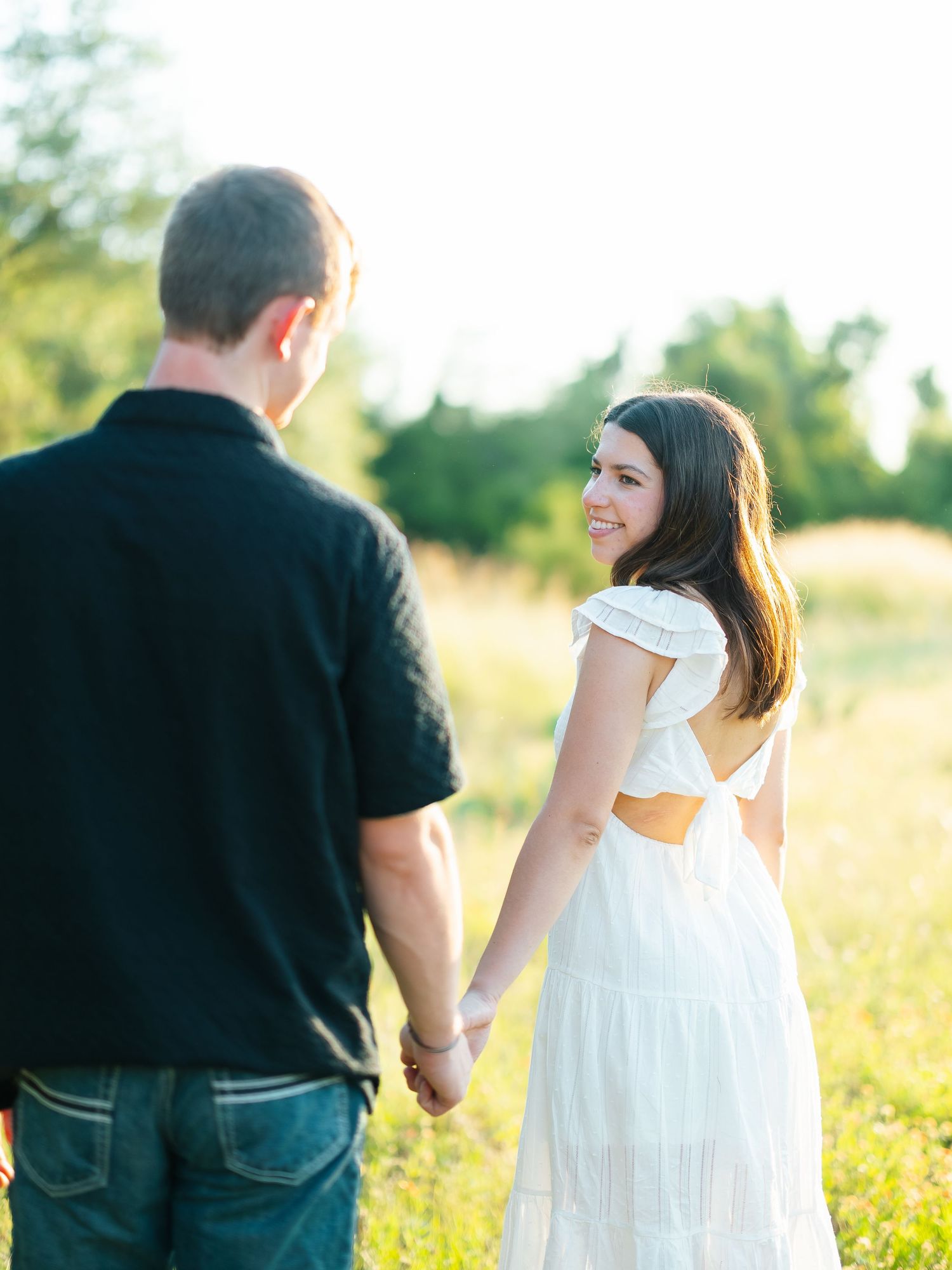 Woman in white backless dress smiles over shoulder while holding hands with partner in sunny field.