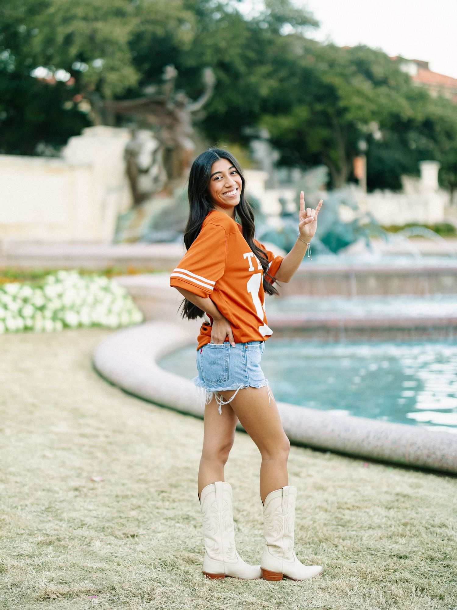 Young woman in orange Texas Longhorns jersey, denim skirt, and white cowboy boots making peace sign by fountain.