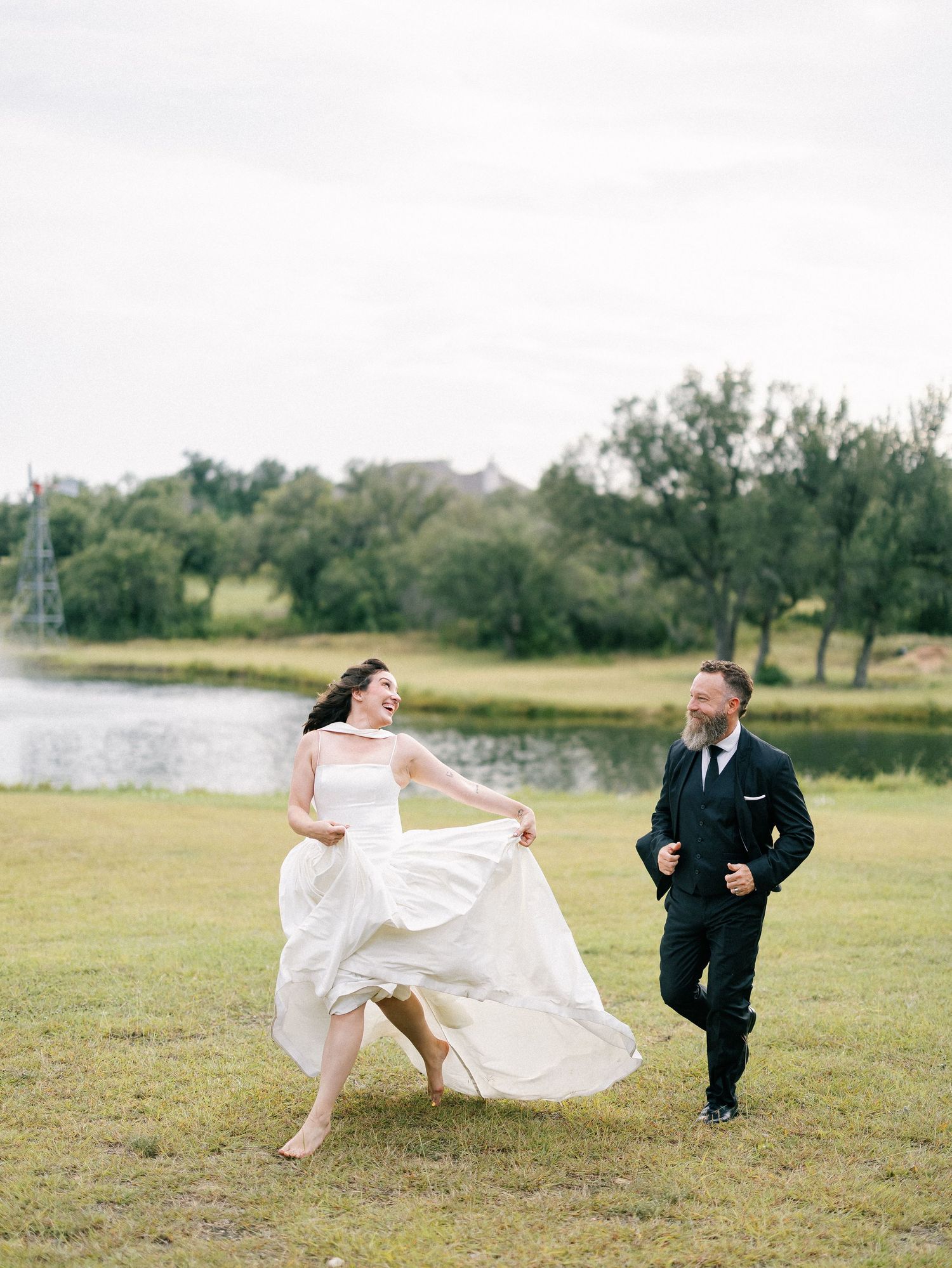 Joyful bride in white gown and groom in black suit running together on green lawn near a lake on their wedding day.