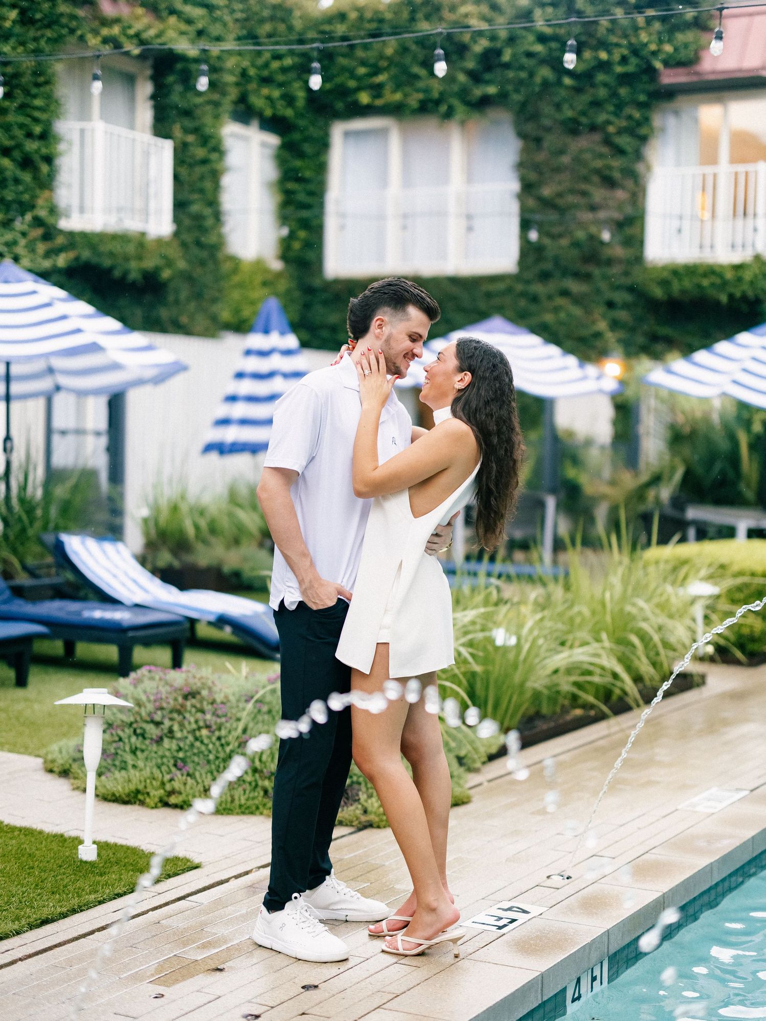 Couple embracing romantically by a pool with blue striped umbrellas and ivy-covered building backdrop.