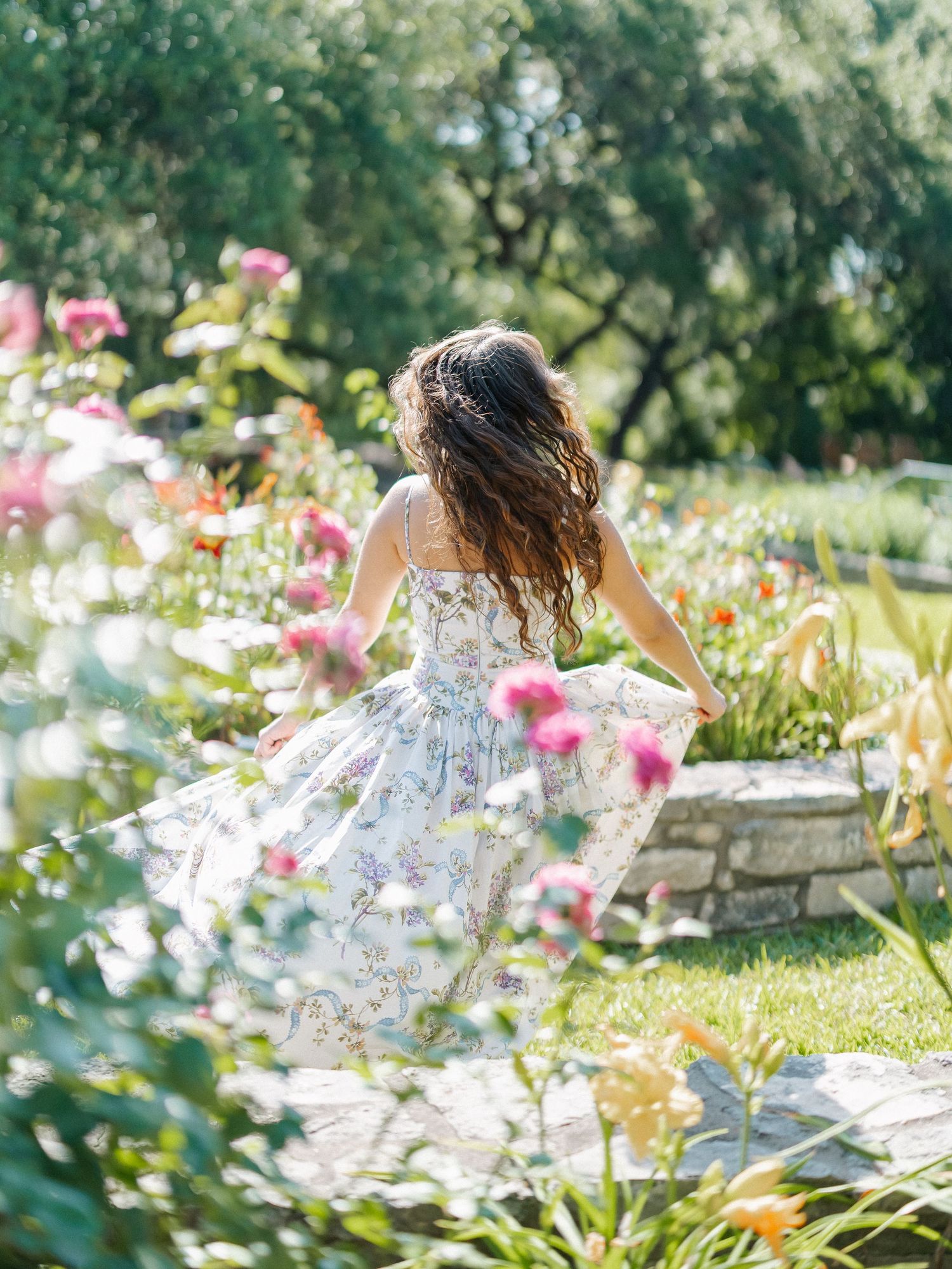 A girl in a floral dress twirls in a sunlit garden blooming with pink and orange flowers.