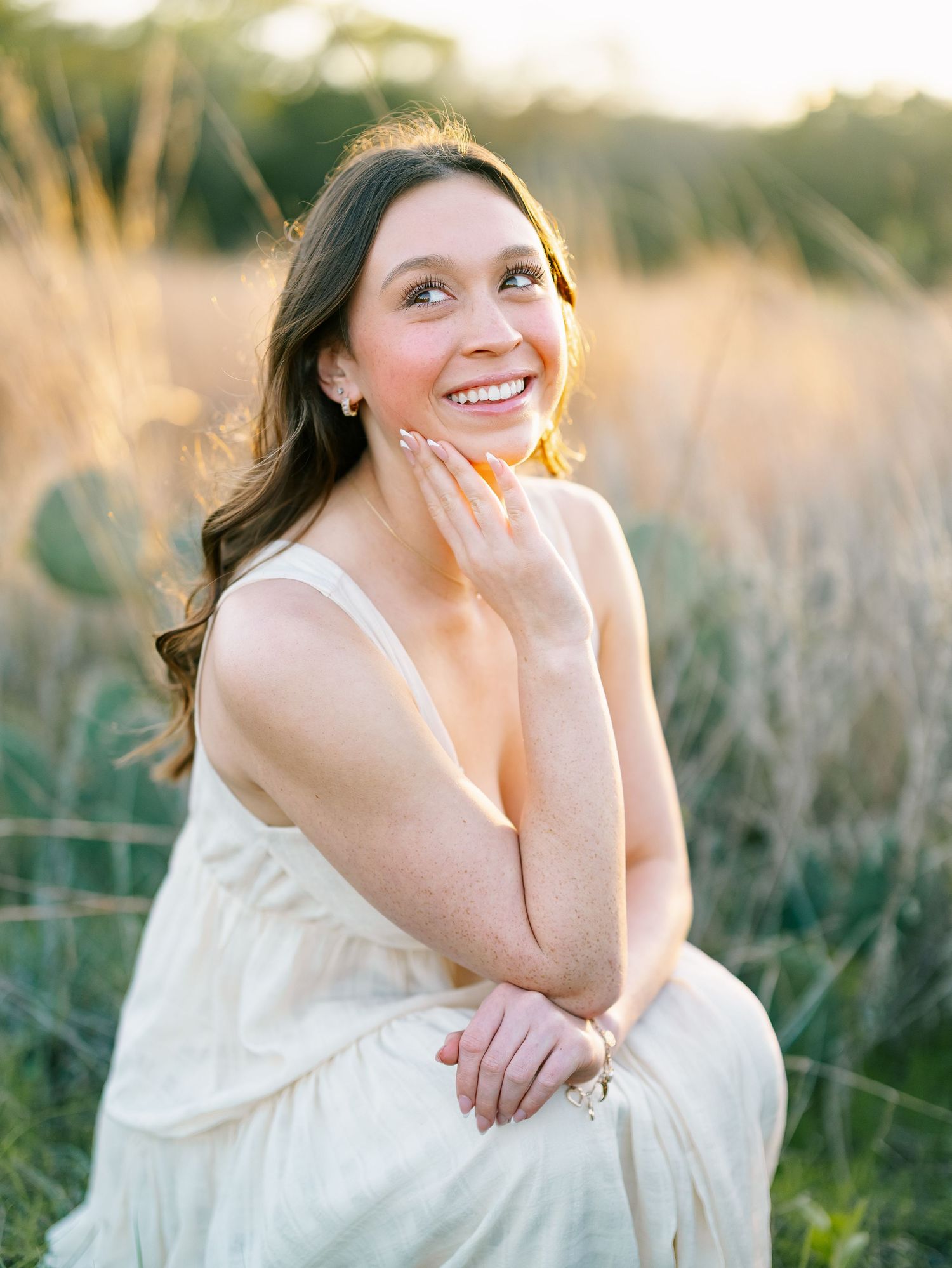 A smiling brunette woman in a cream dress sits gracefully in a golden field at sunset.