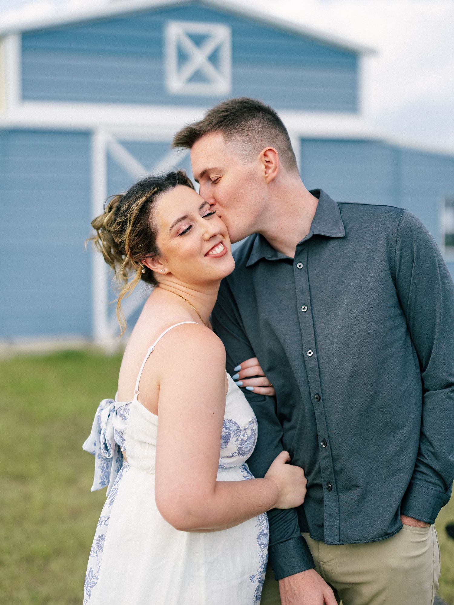 Couple embracing in front of a blue barn, man kissing woman on cheek as she smiles and sticks out her tongue.