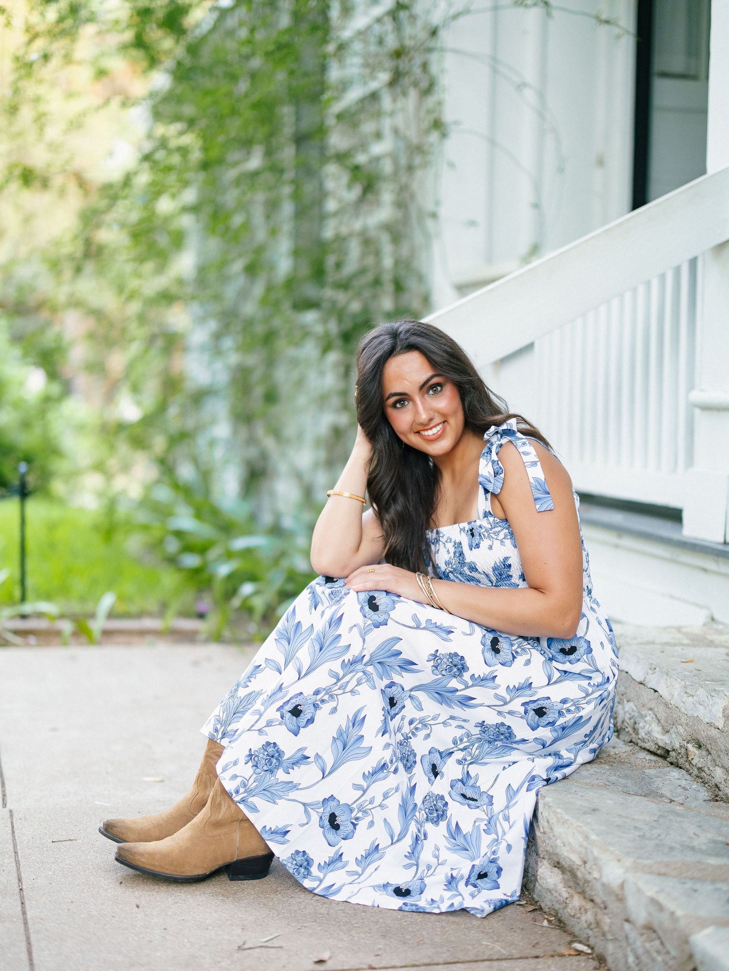 A young woman in a blue floral dress and tan boots sits on stone steps beside a white building.