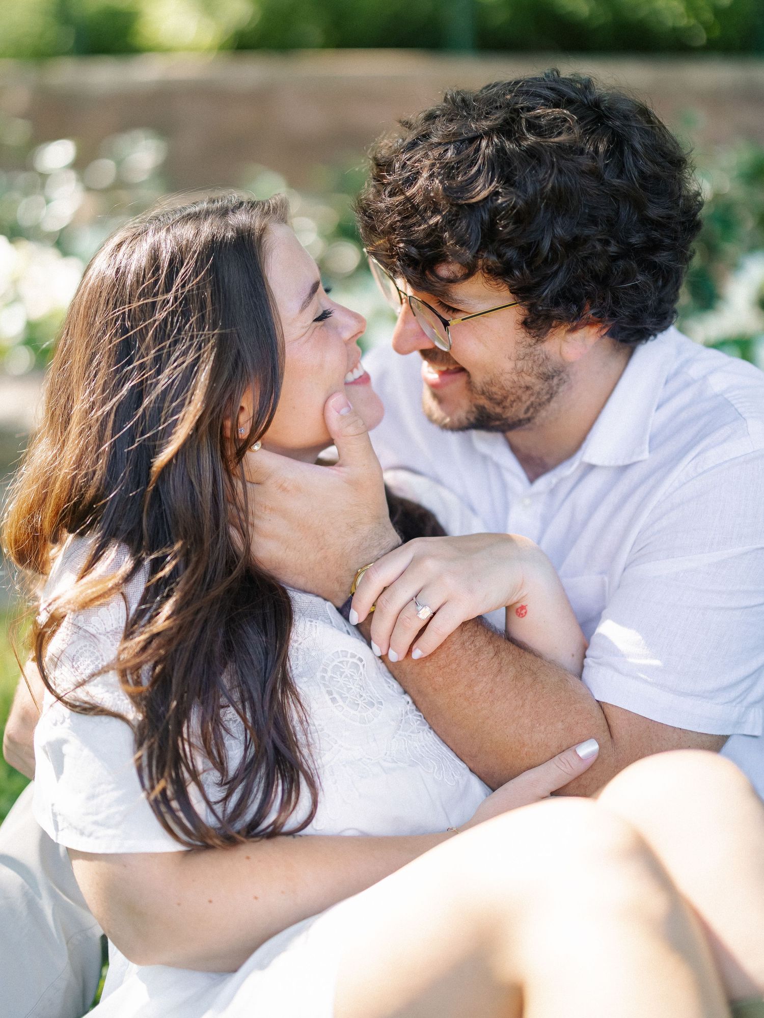 A couple gazes lovingly at each other while sitting outdoors, the woman wearing a white dress with an engagement ring.