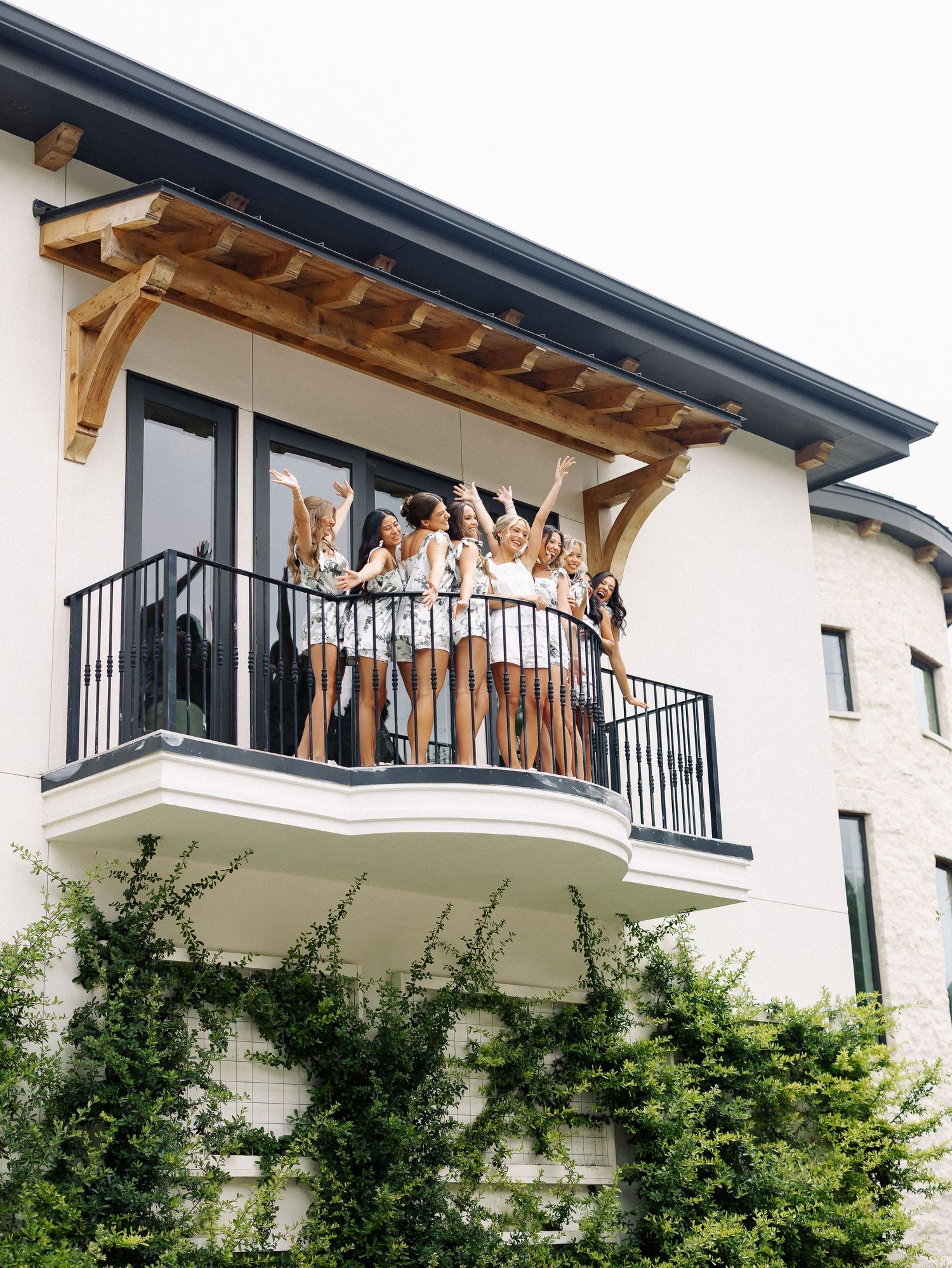 Group of women celebrating on a balcony of a modern white brick building with wooden pergola overhead.