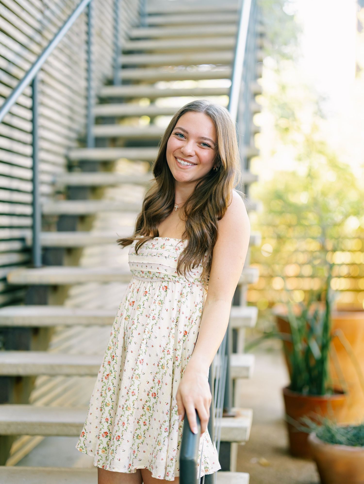 Brunette woman in floral sundress smiling on outdoor staircase with greenery background.