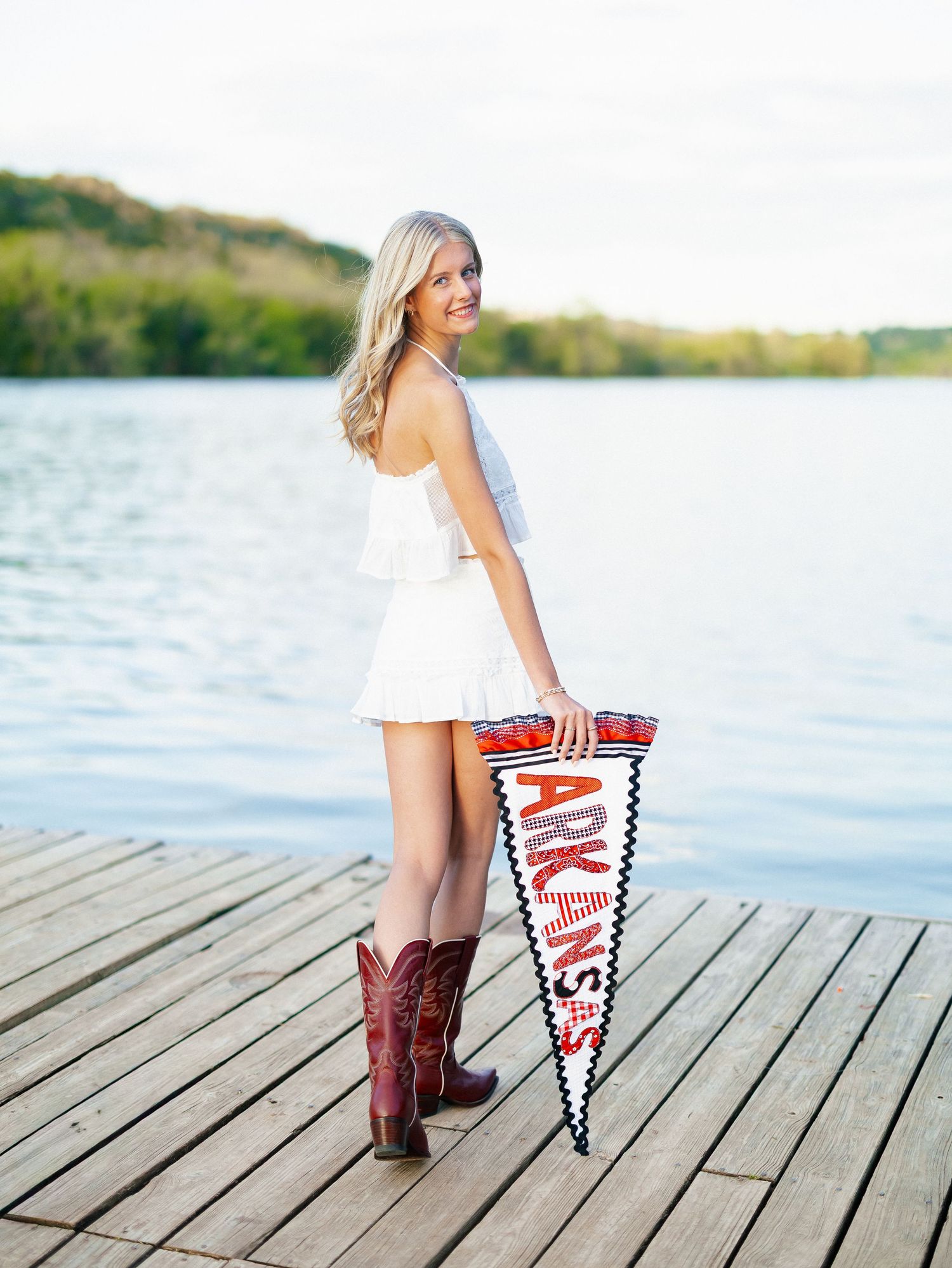 A blonde young woman in a white dress and cowboy boots holds an Arkansas pennant on a lake dock.
