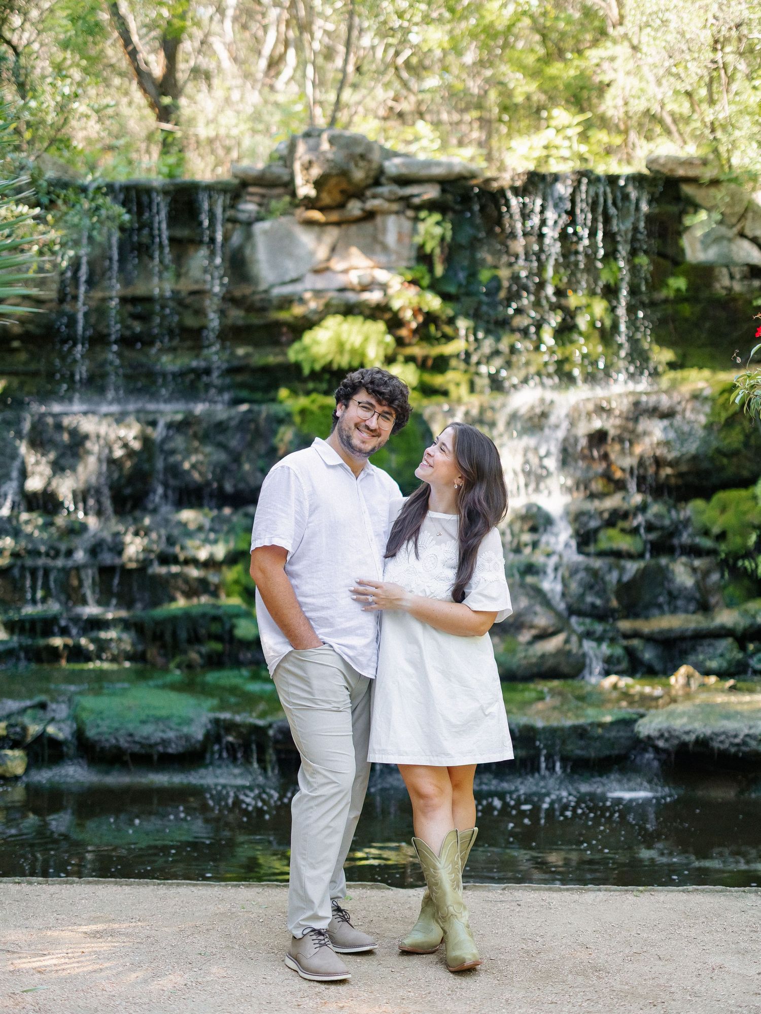 Couple posing together in front of a scenic waterfall surrounded by lush greenery, wearing coordinating white outfits.