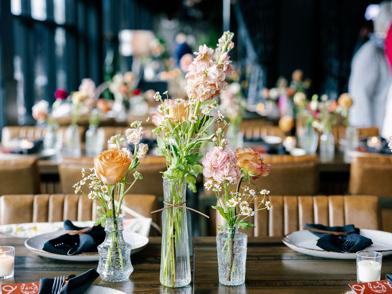 Elegant wedding reception table with glass vases of pink and peach flowers, black napkins, and white plates in a warmly lit venue.