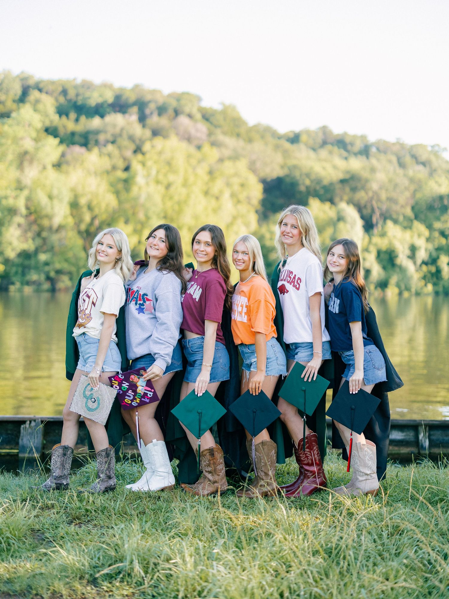 Six young women in college sweatshirts and cowboy boots pose with graduation caps by a lake.