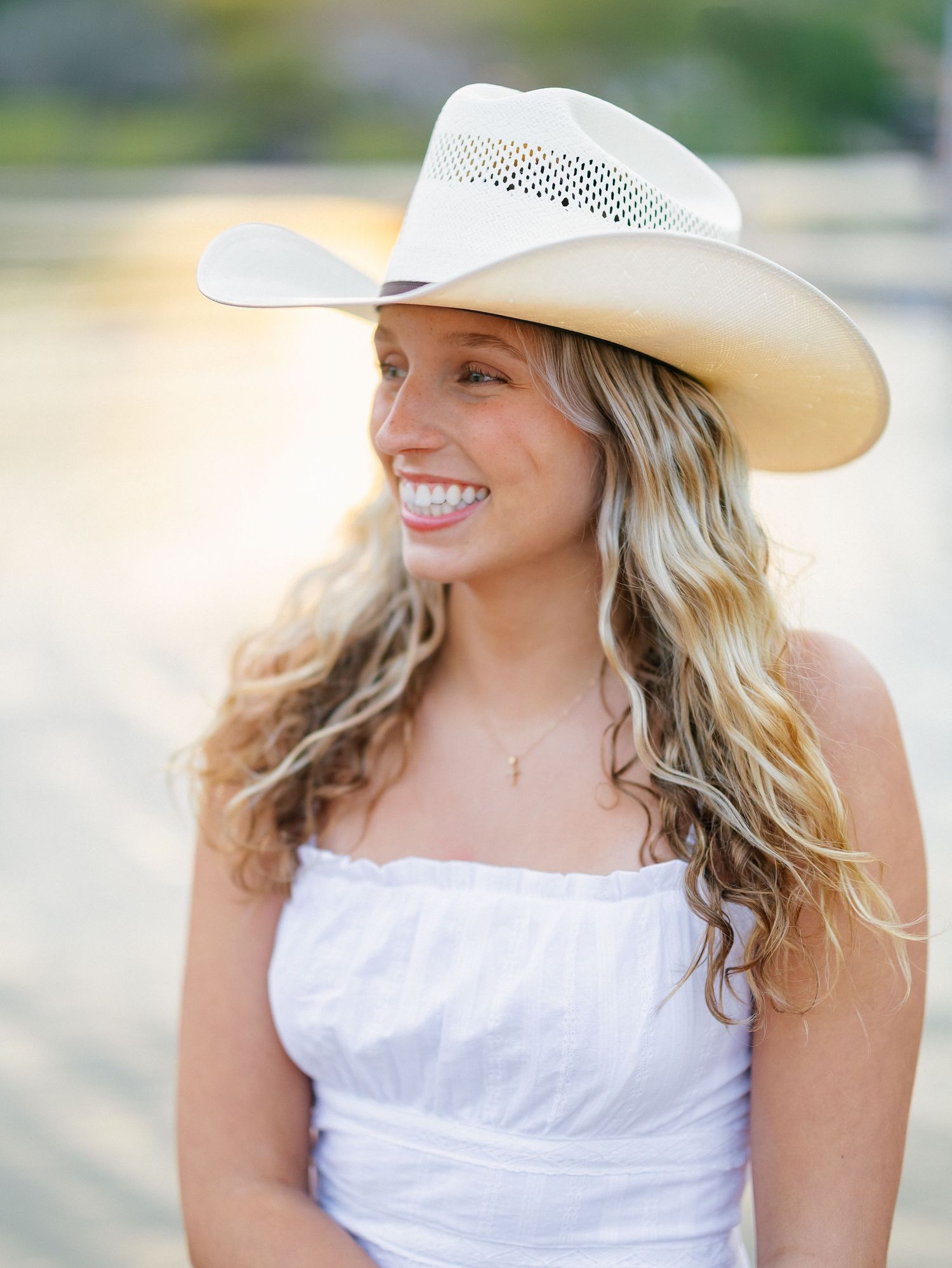 Smiling young woman with wavy blonde hair wearing a white cowboy hat and white strapless top outdoors.