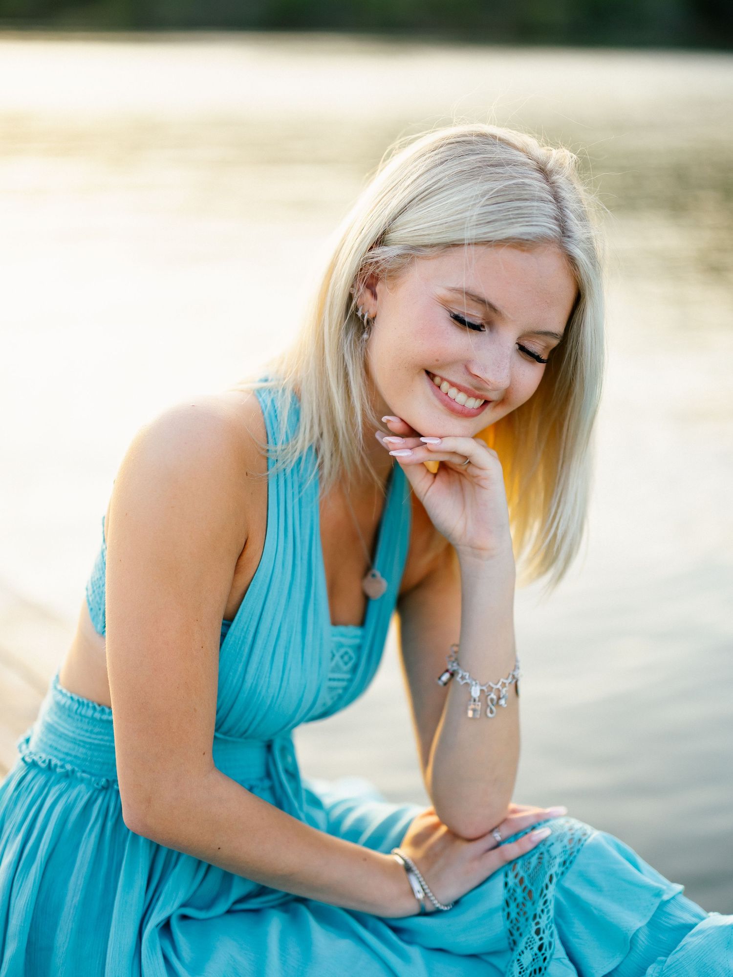 Smiling blonde woman in turquoise dress sitting by water with silver bracelet jewelry.