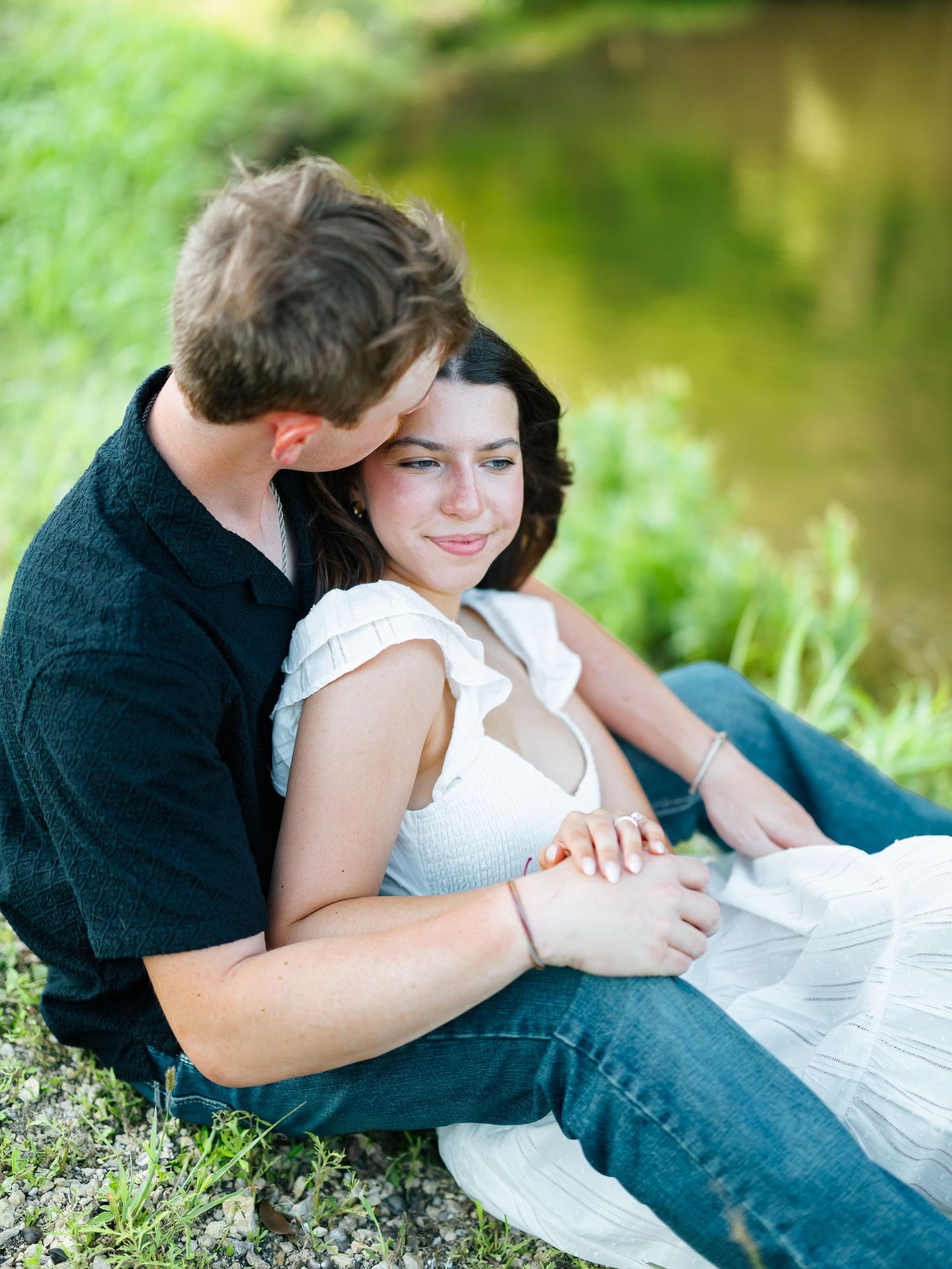 Couple sitting by a pond, man kissing woman's head while she smiles softly in a white dress and jeans.
