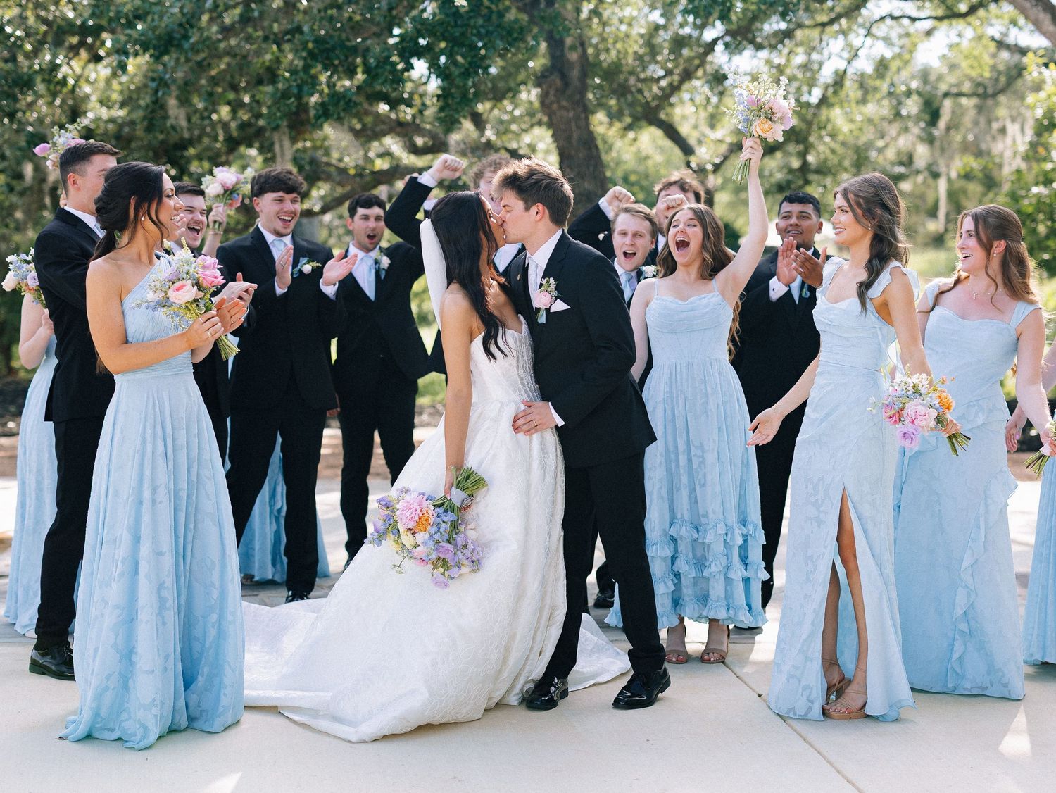 Wedding party poses outdoors, bride and groom kissing, bridesmaids in light blue gowns, groomsmen in black suits.