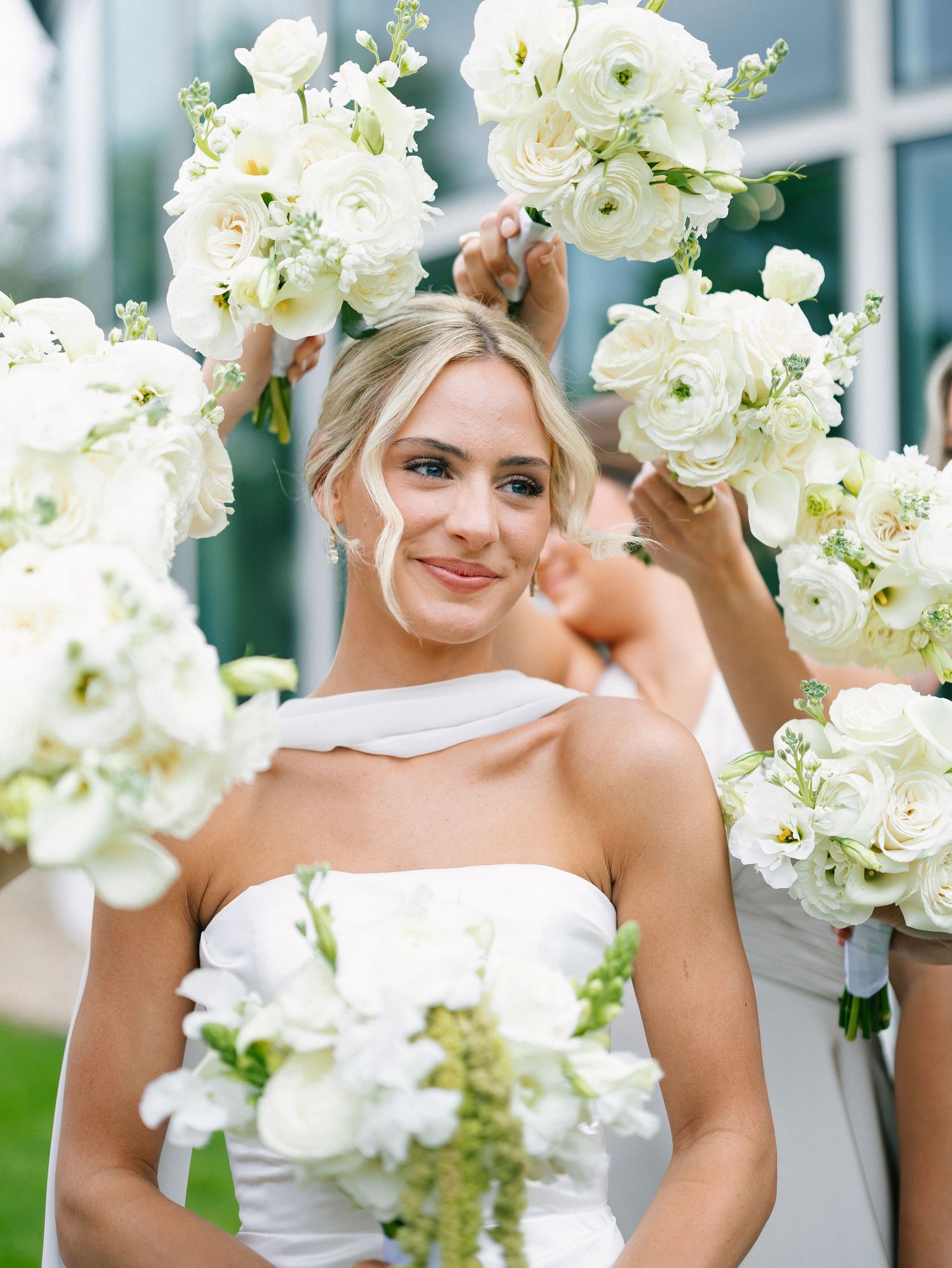 Blonde bride in white strapless gown surrounded by white floral bouquets held by bridesmaids.