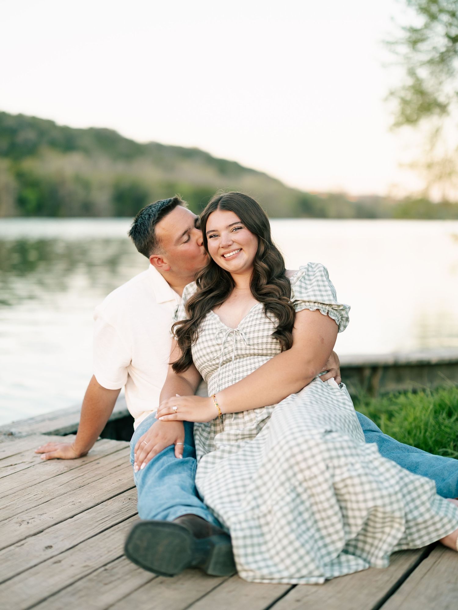 Happy couple sitting on wooden dock by lake sharing a kiss during engagement session.