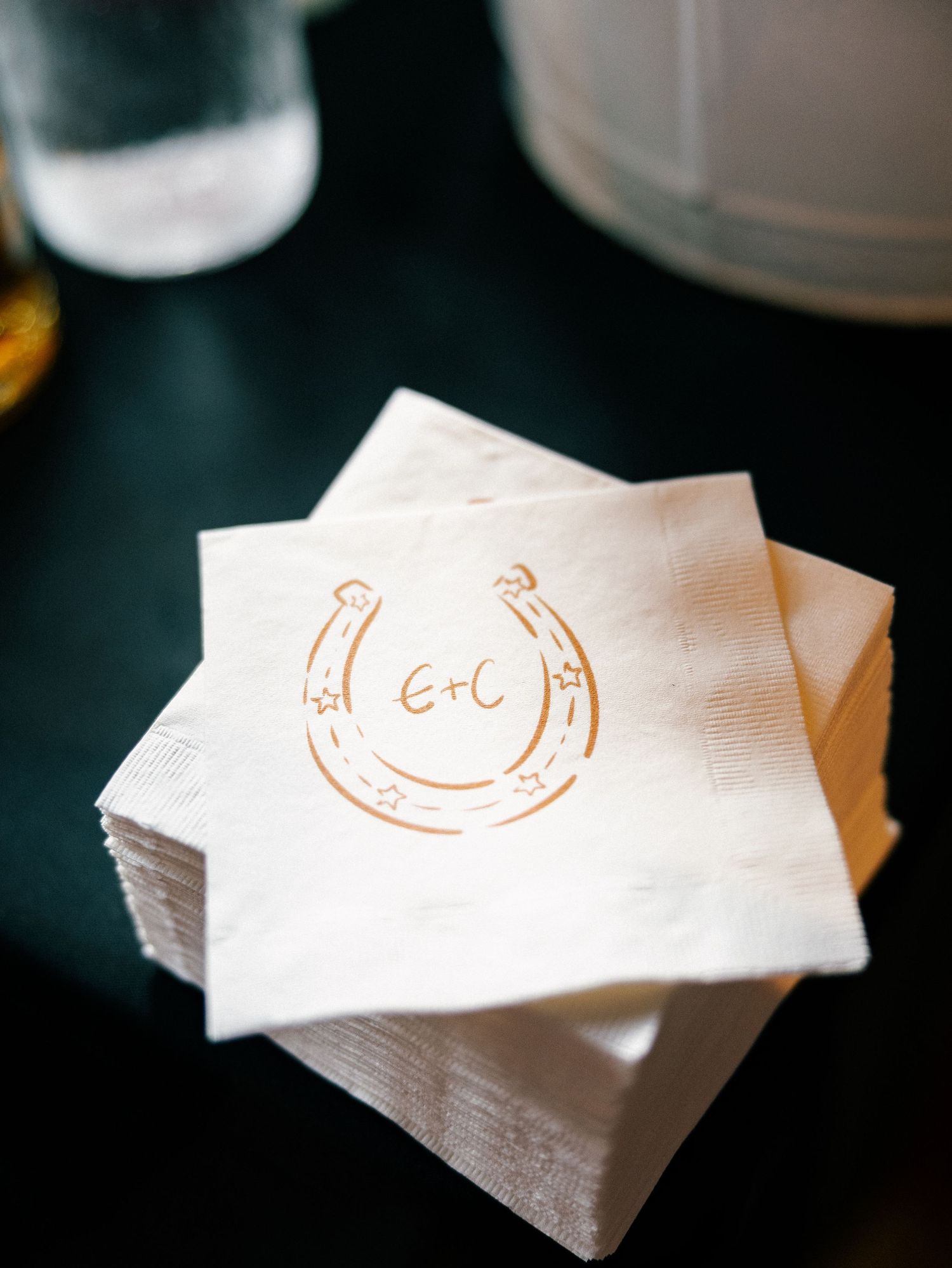 Custom white wedding napkins printed with a horseshoe monogram and initials E+C in gold ink, stacked on a dark surface.