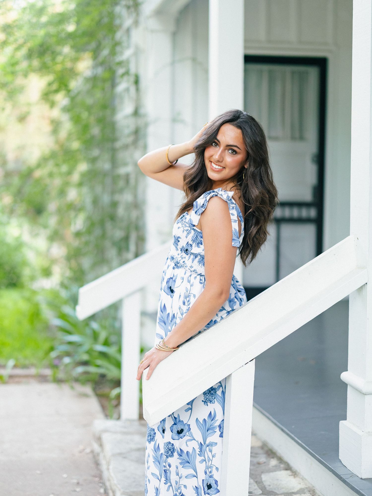 Woman in blue floral maxi dress posing on white porch steps, smiling over shoulder in lush garden setting.