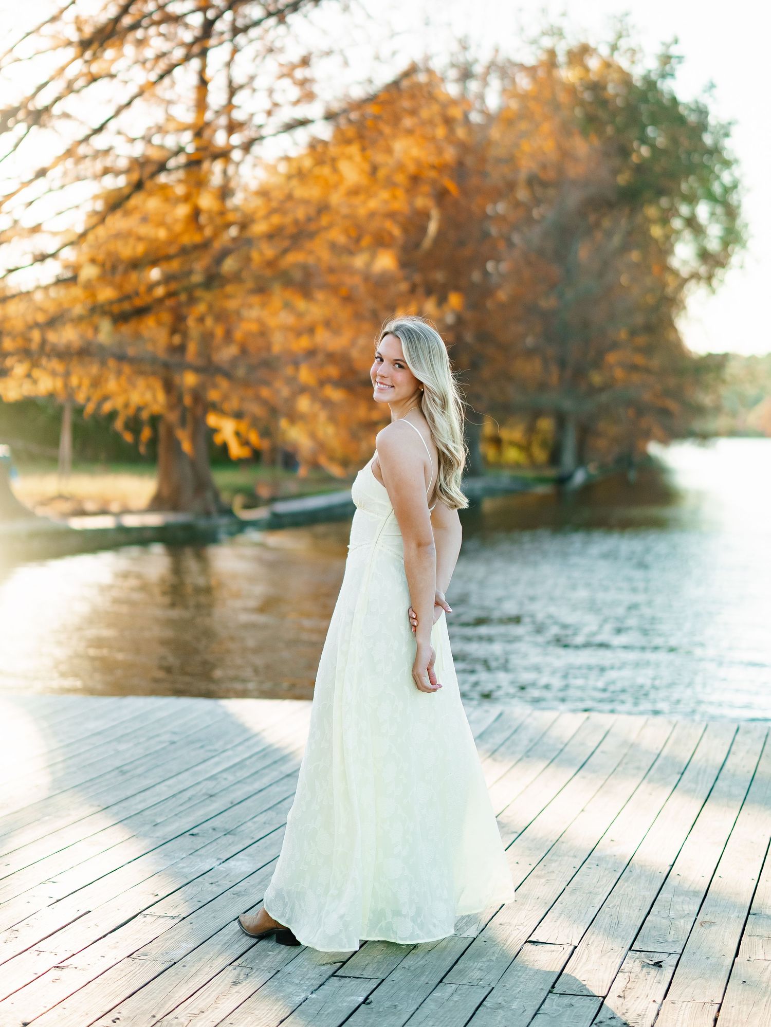 Bride in white gown smiling on wooden dock with autumn foliage and lake backdrop.