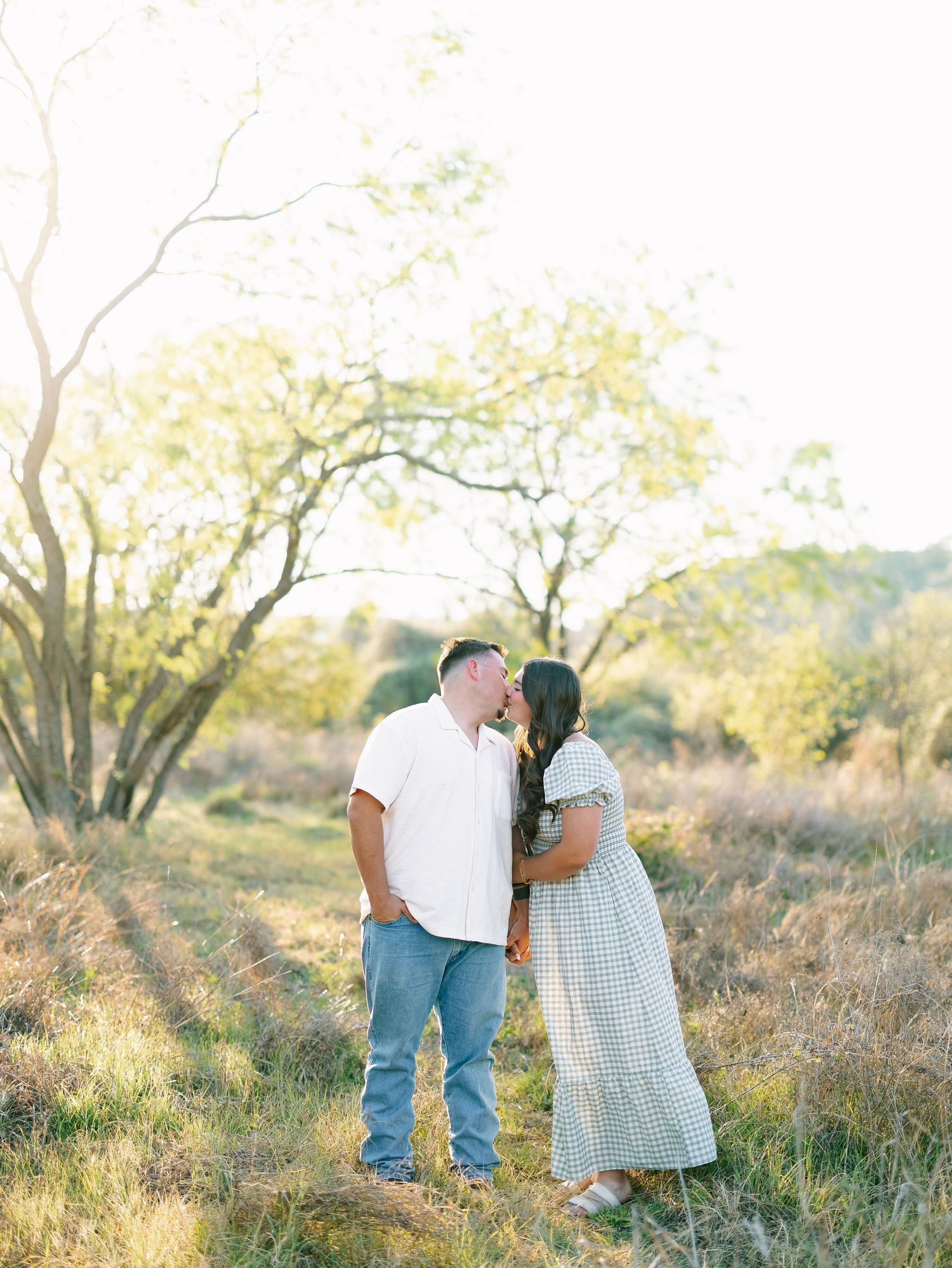 Couple sharing a kiss in golden sunlit field during outdoor engagement photo session.