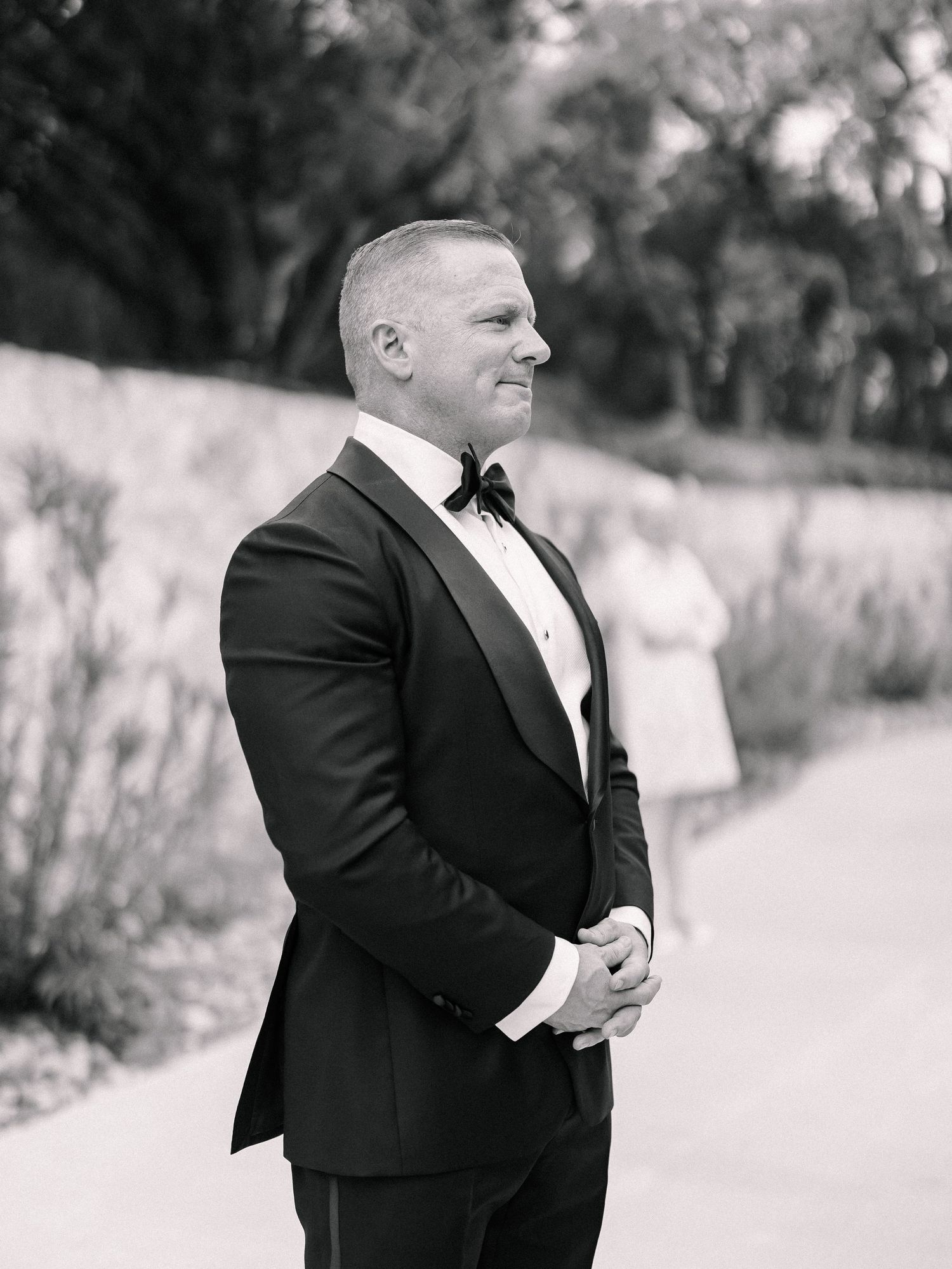Groom in classic black tuxedo and bow tie smiles while waiting outdoors during wedding day first look.