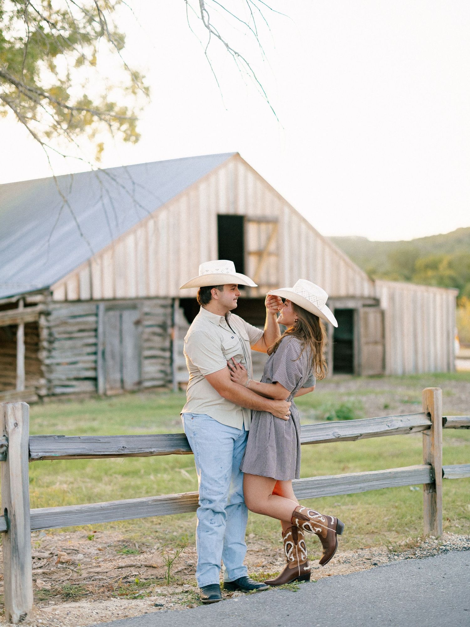 Couple in cowboy hats embracing near rustic barn fence in golden hour light, wearing boots and casual country attire.