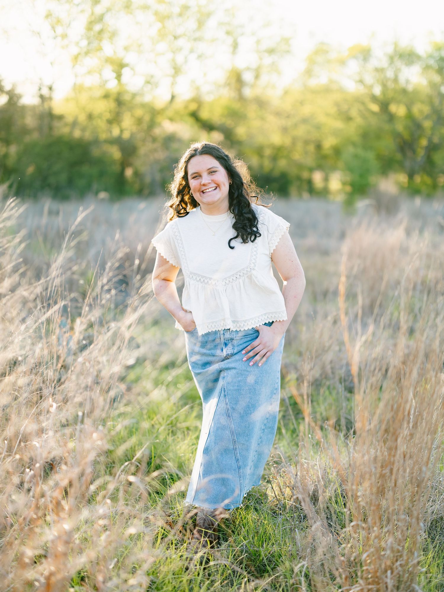 A smiling young woman in a white top and jeans poses in a sunlit field with tall grass and green trees.