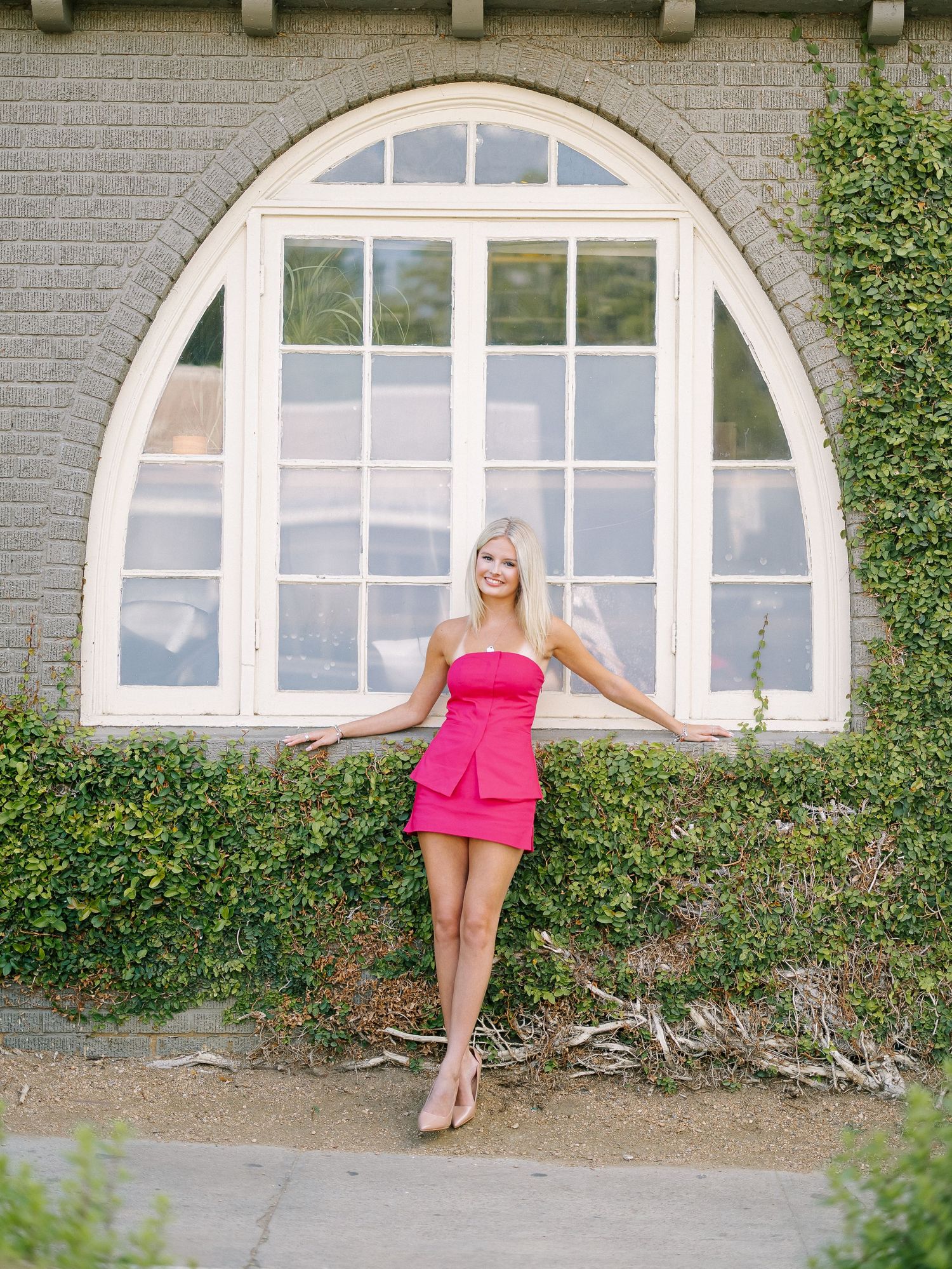 Blonde woman in pink strapless mini dress posing in front of arched window covered in ivy on brick building.