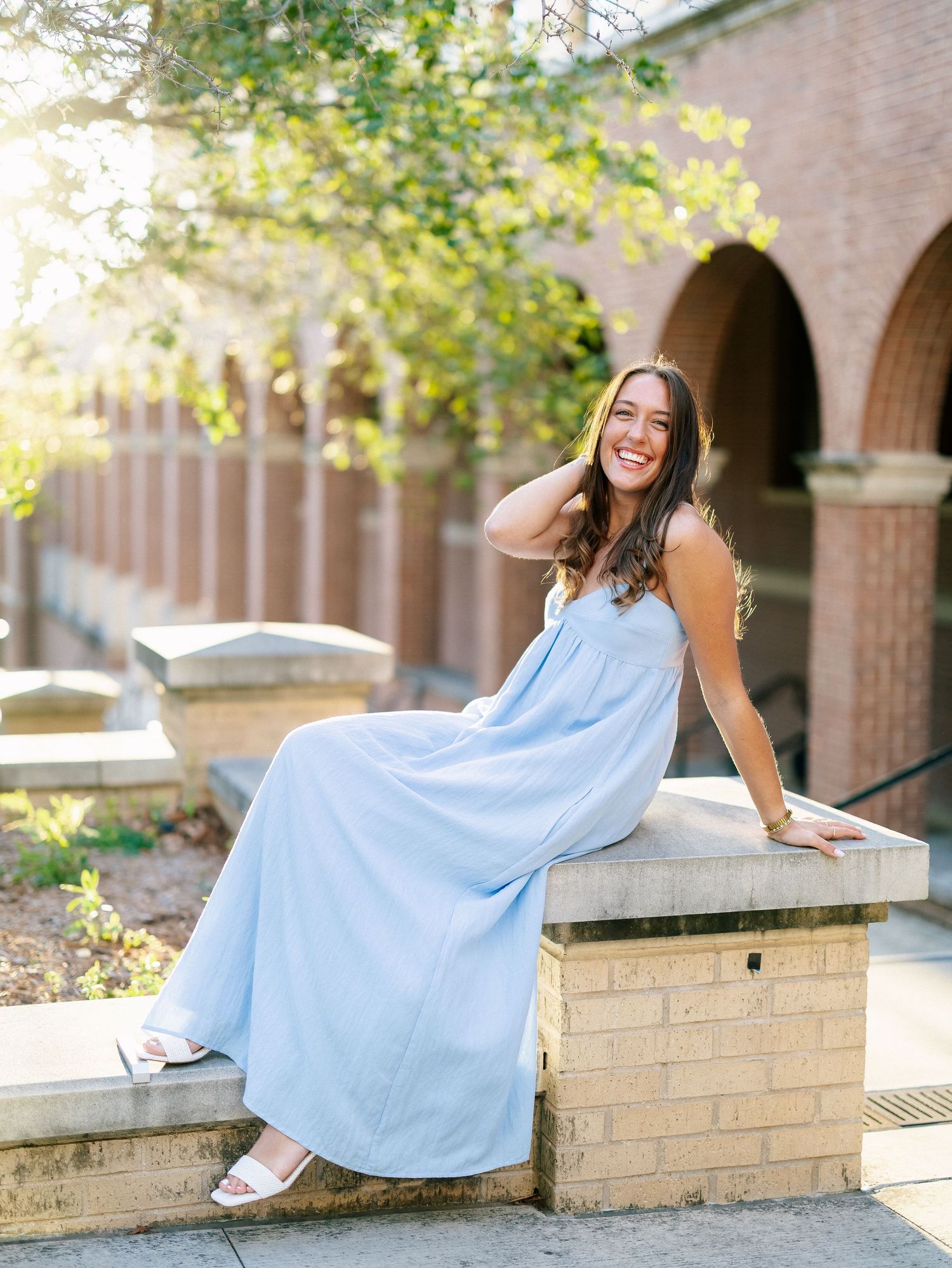 A young woman in a light blue maxi dress sits smiling on a brick wall outside a collegiate building.
