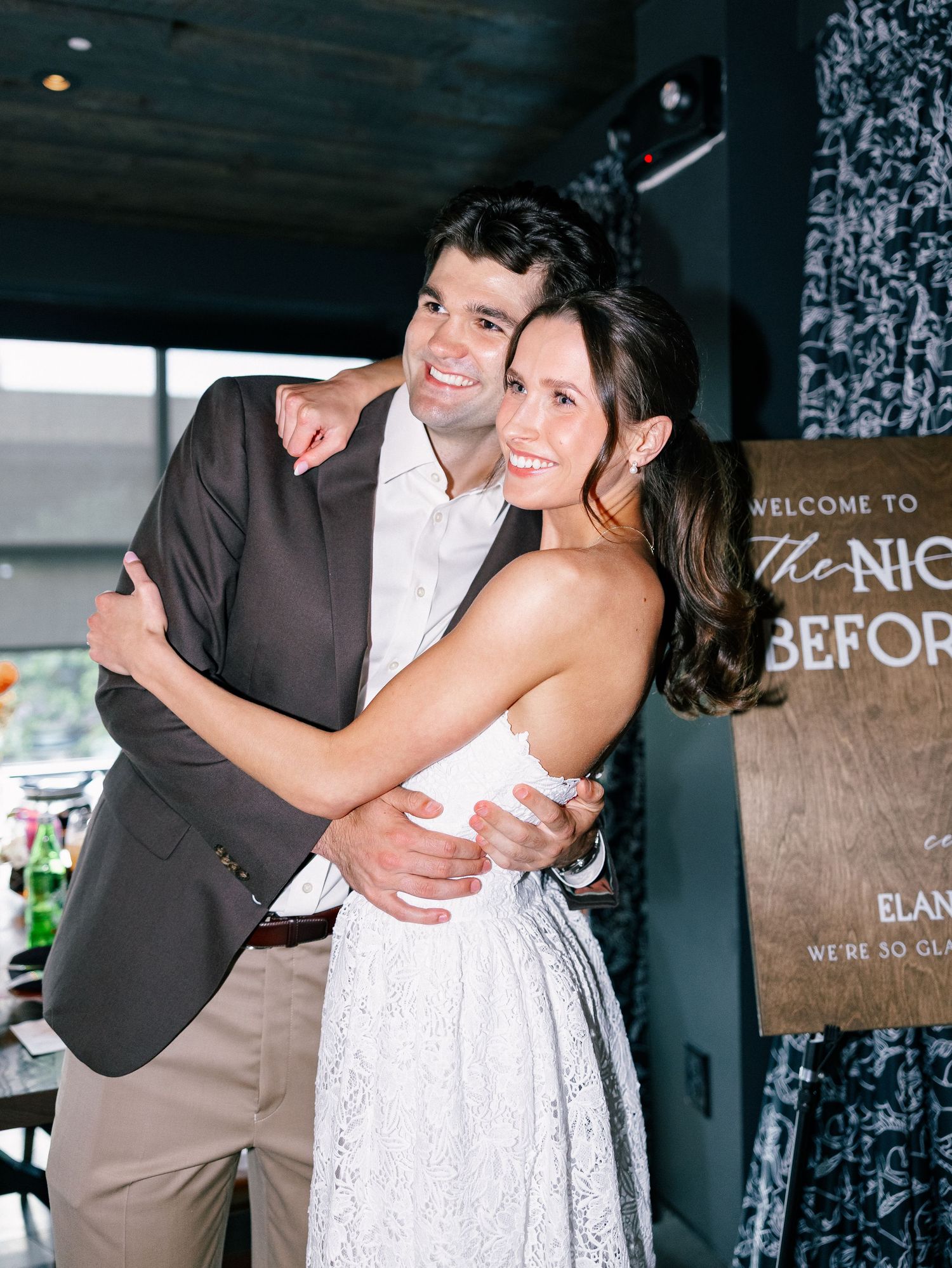 Smiling couple embraces at a wedding eve party in front of a welcome sign.