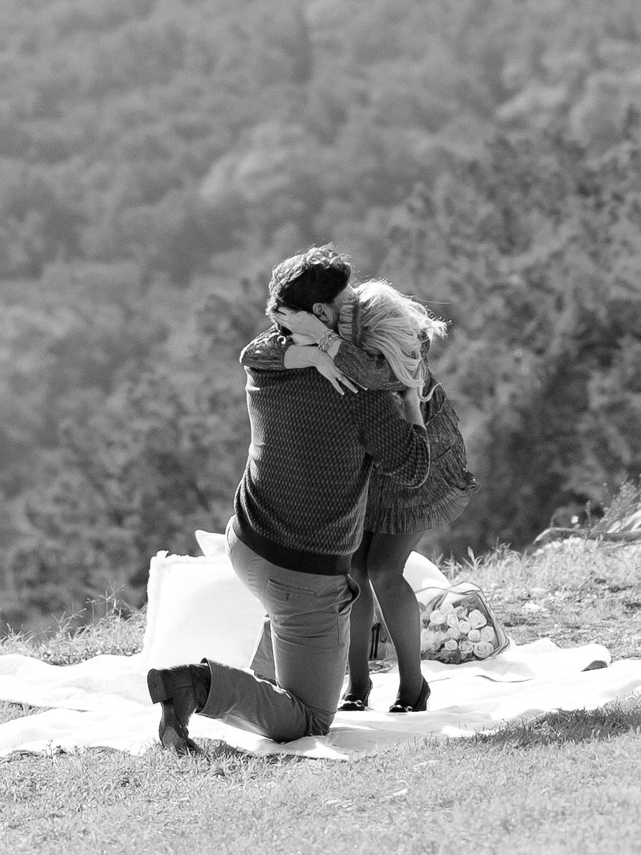 Black and white photo of a man on one knee proposing to his partner on a hillside blanket.