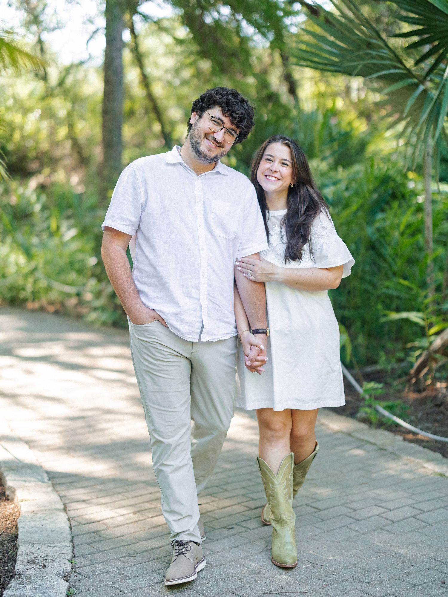 A smiling couple walks hand-in-hand along a garden path, dressed in white outfits with the woman wearing green cowboy boots.