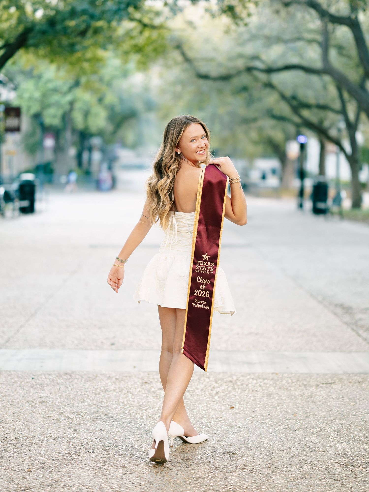 Blonde woman in white dress walking away showing Texas A&M Class of 2024 graduation stole.