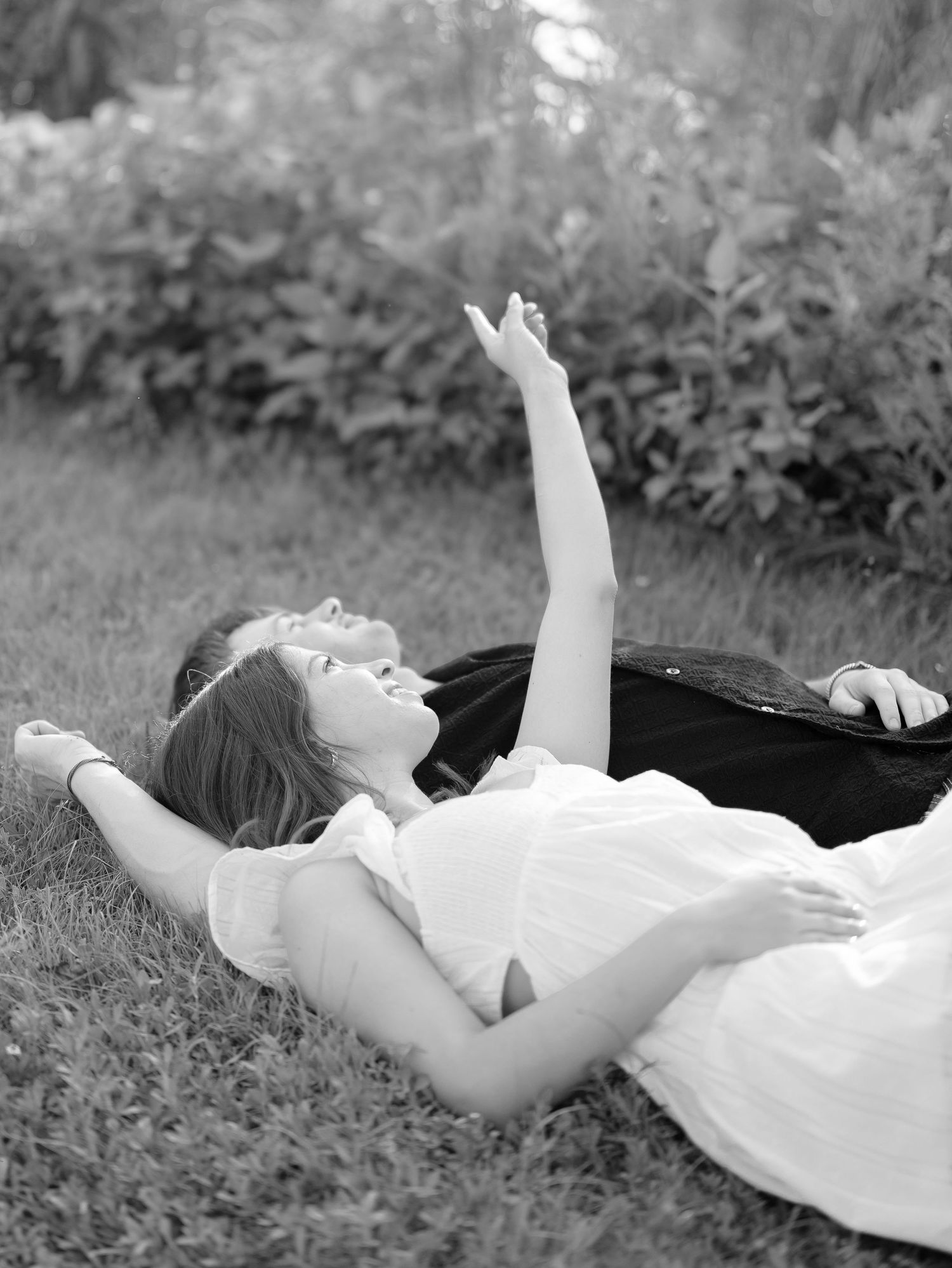 Black and white photo of young couple lying on grass laughing with arm raised toward sky.