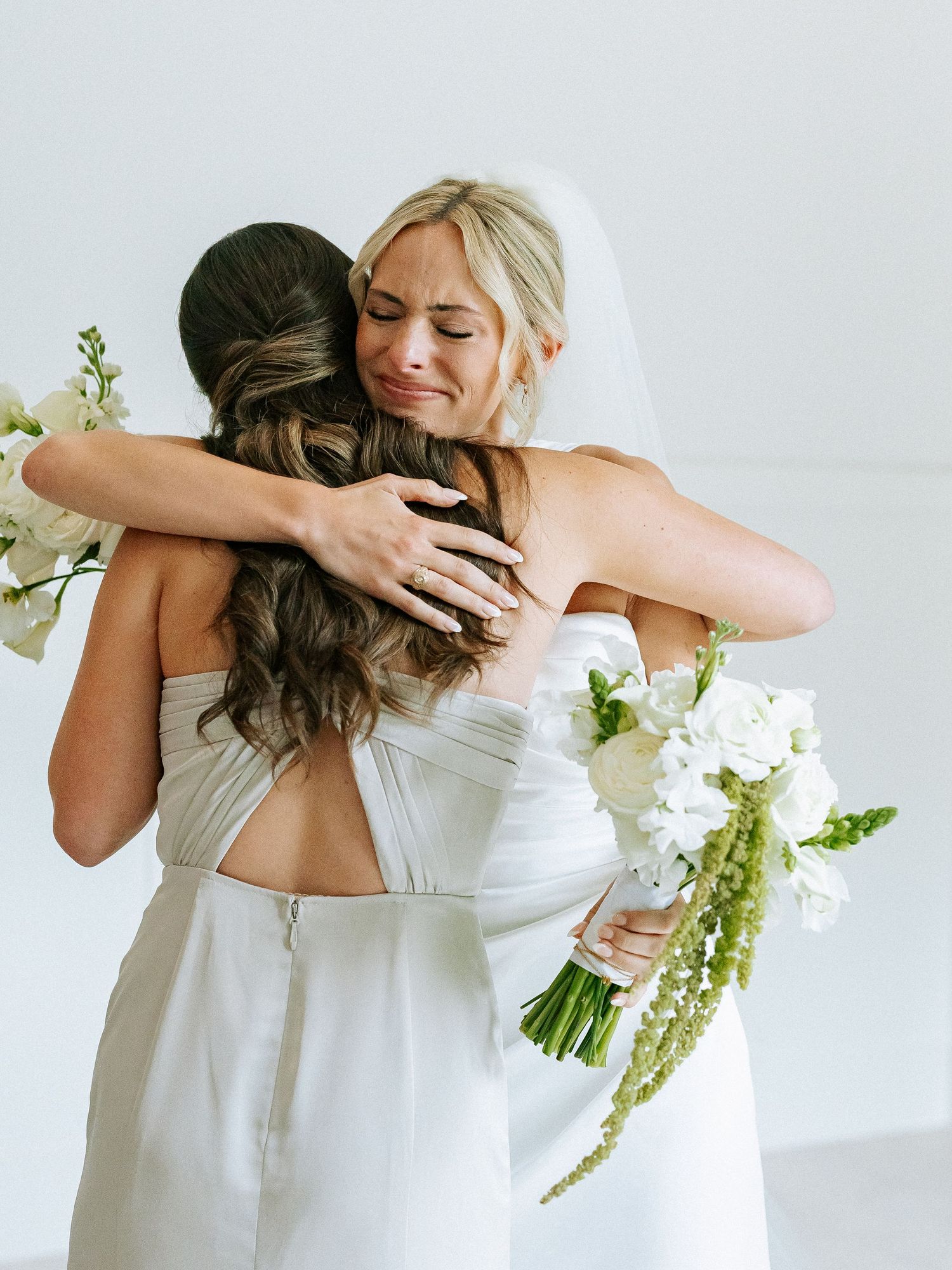 Two brides share an emotional hug on wedding day, one crying while holding a white floral bouquet.