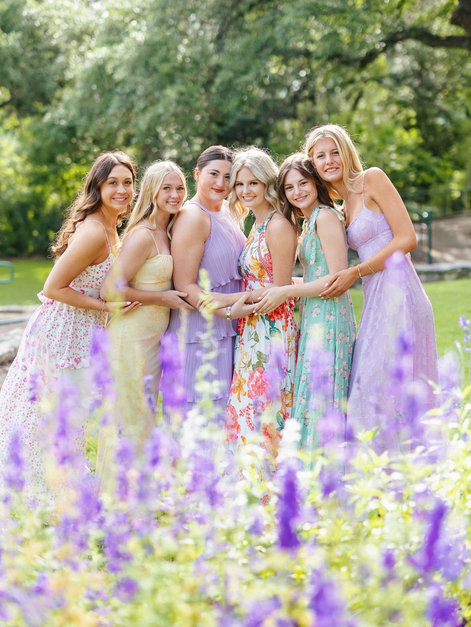 Six women in pastel dresses pose together smiling in a garden with purple wildflowers in the foreground.