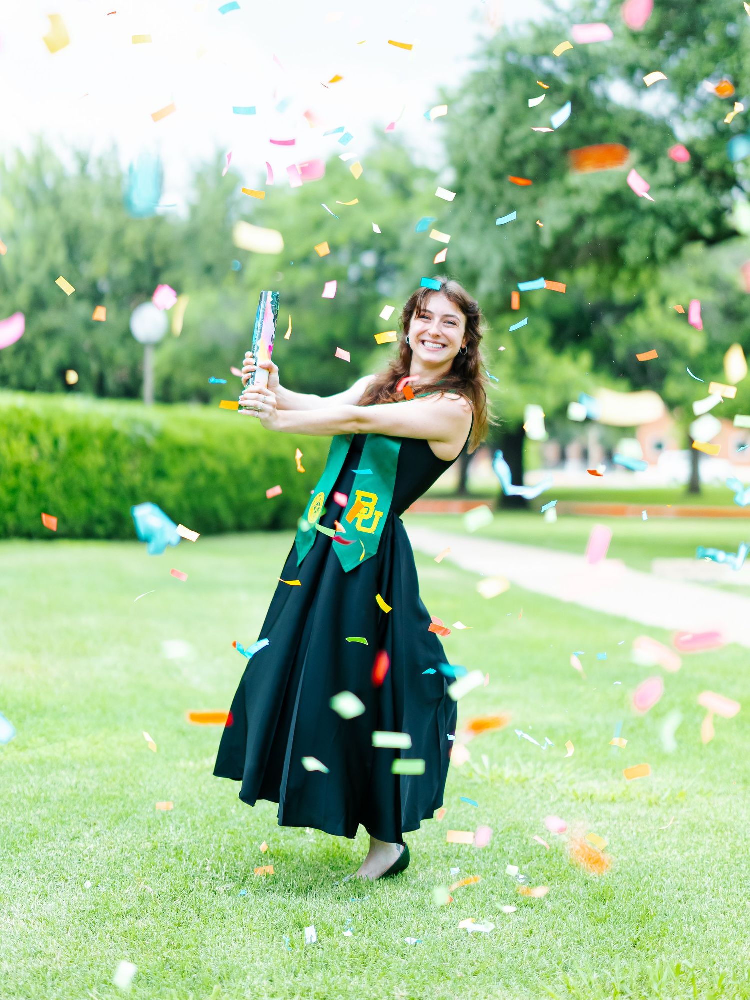 Smiling graduate in black dress and green sash shoots confetti cannon outdoors on green lawn during celebration.