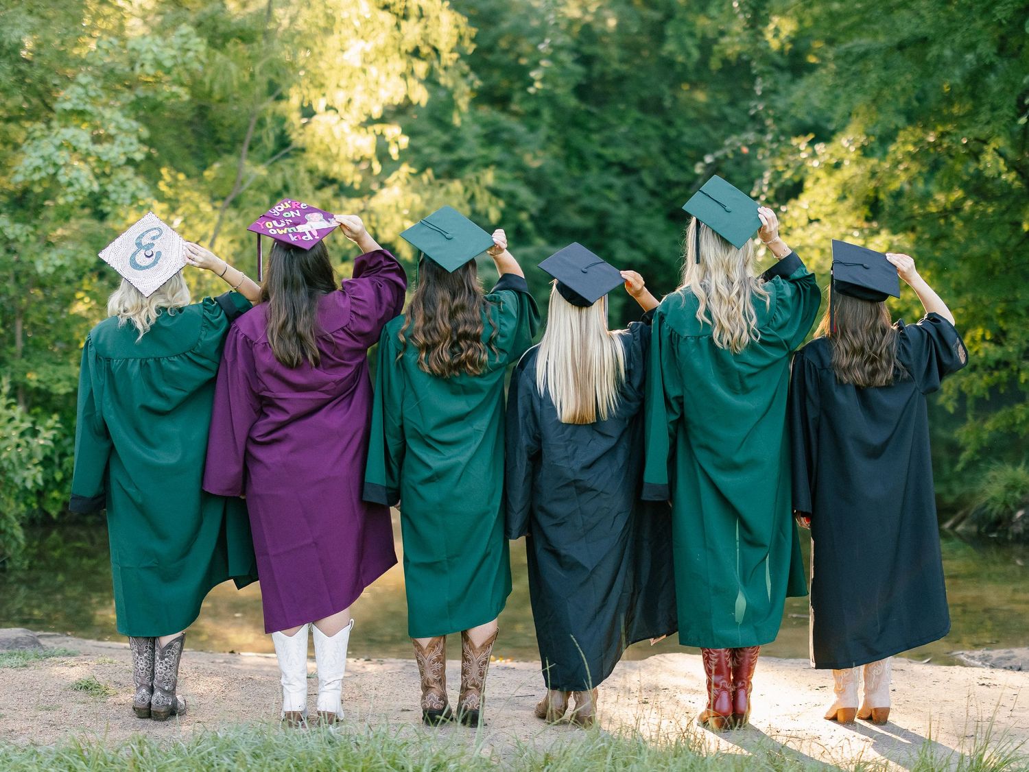 Seven female graduates in green caps and gowns pose with backs to camera outdoors among lush green trees.