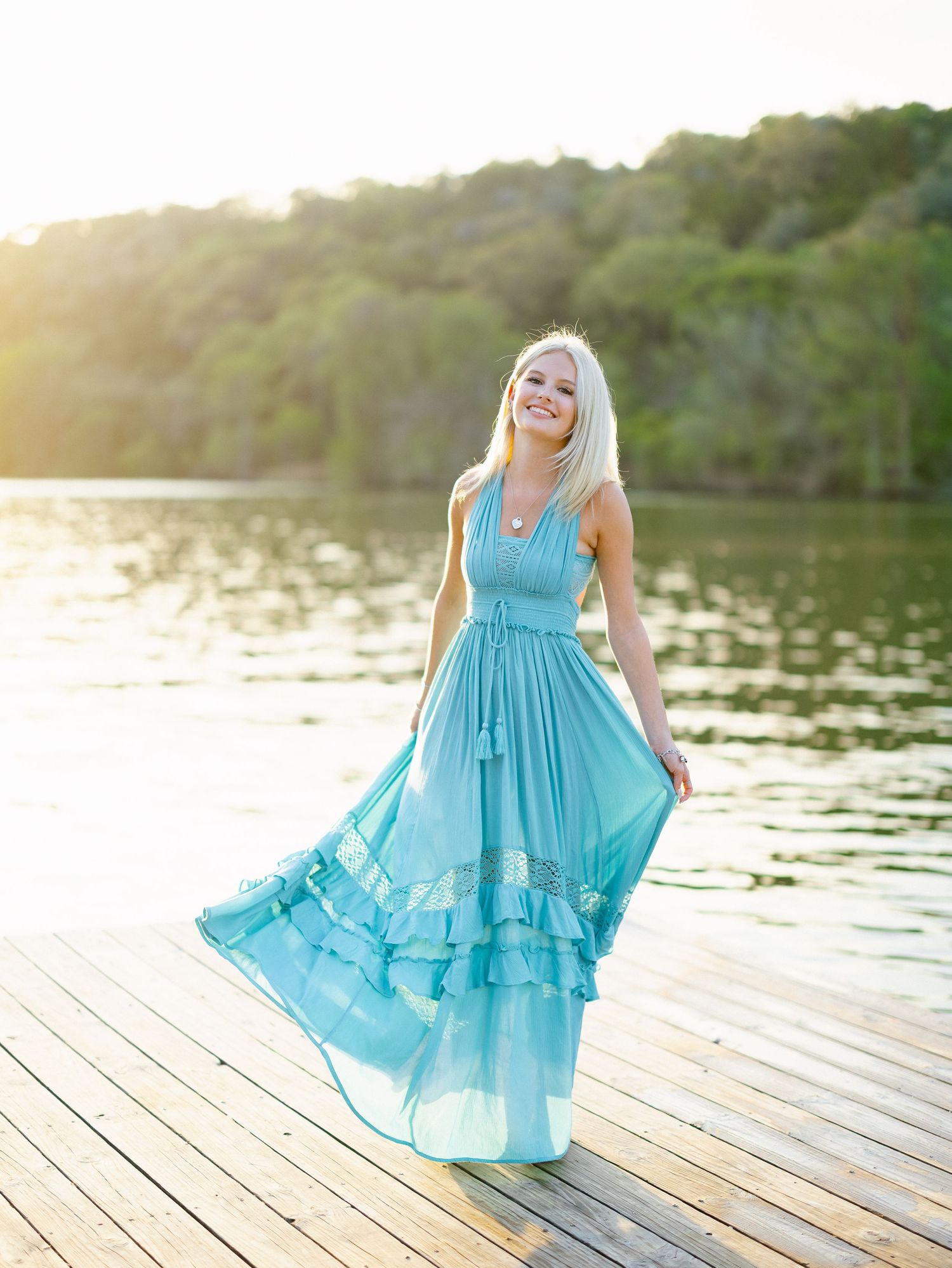 Smiling blonde senior poses in a teal ruffle maxi dress on a wooden dock by a sunlit lake.